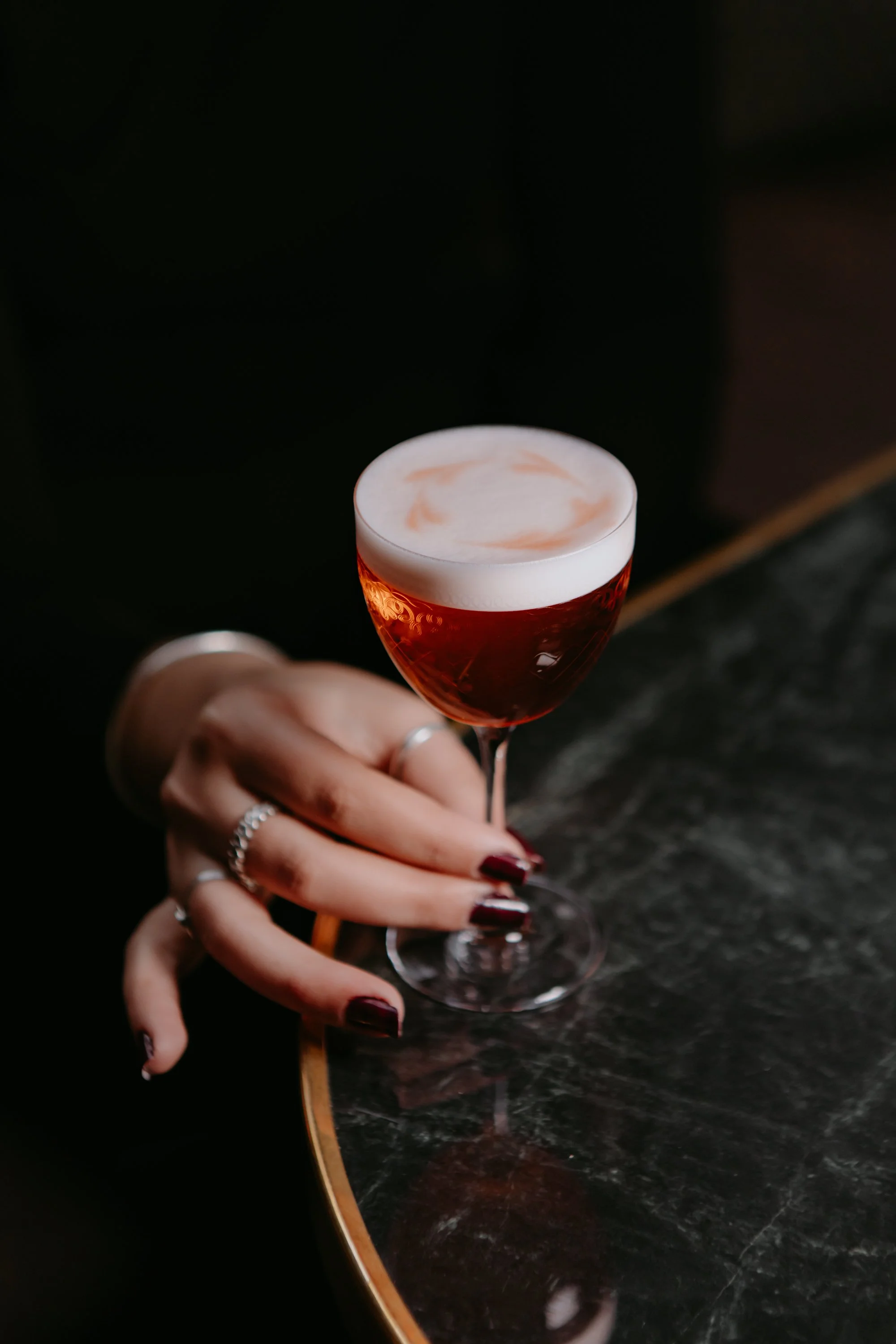Une main tenant un verre à cocktail rouge avec une mousse blanche sur le dessus, placé sur un comptoir en marbre noir.