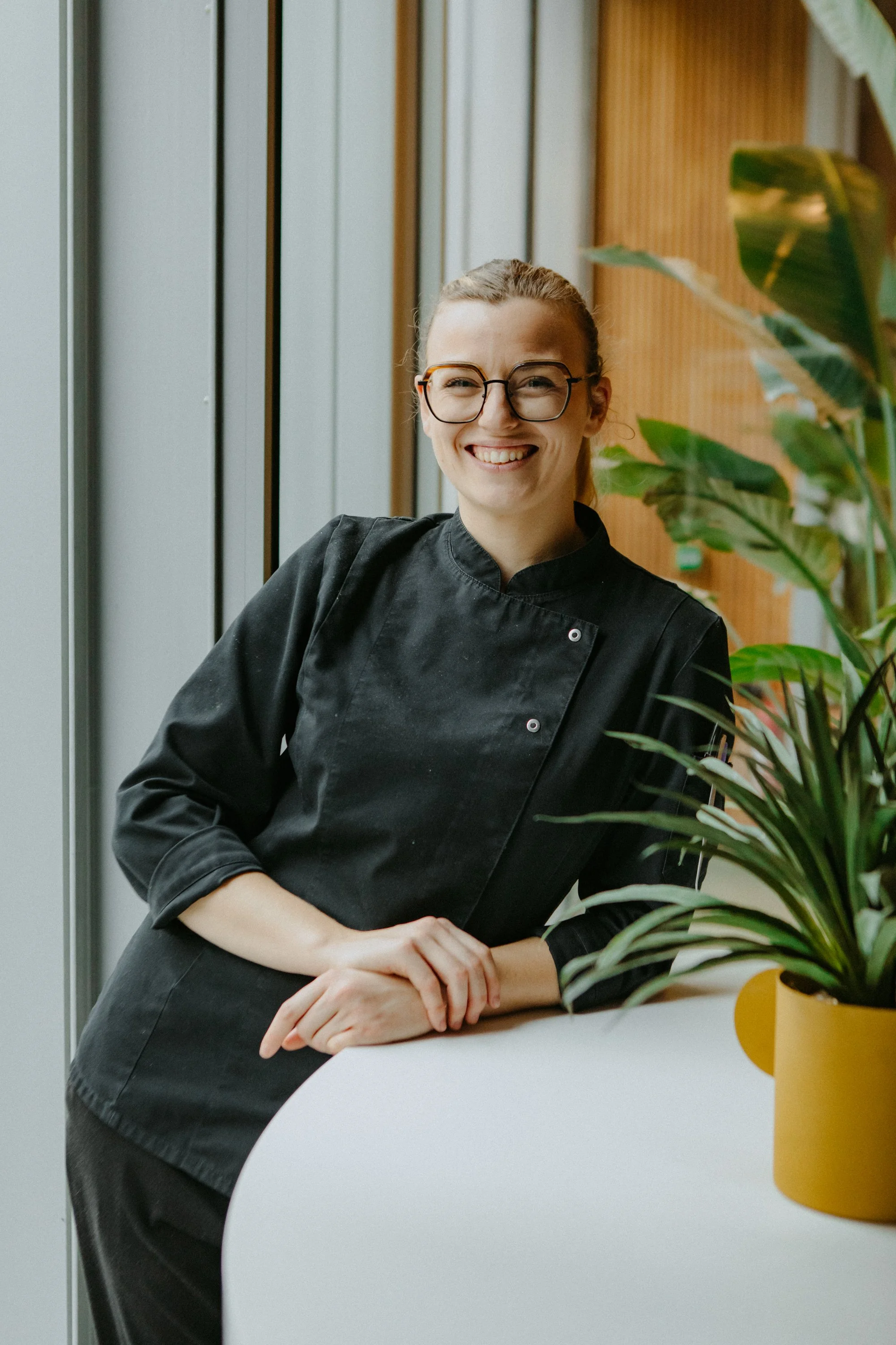 Une femme souriante portant un uniforme de chef cuisinier, posant près d'une plante verte dans un espace moderne.