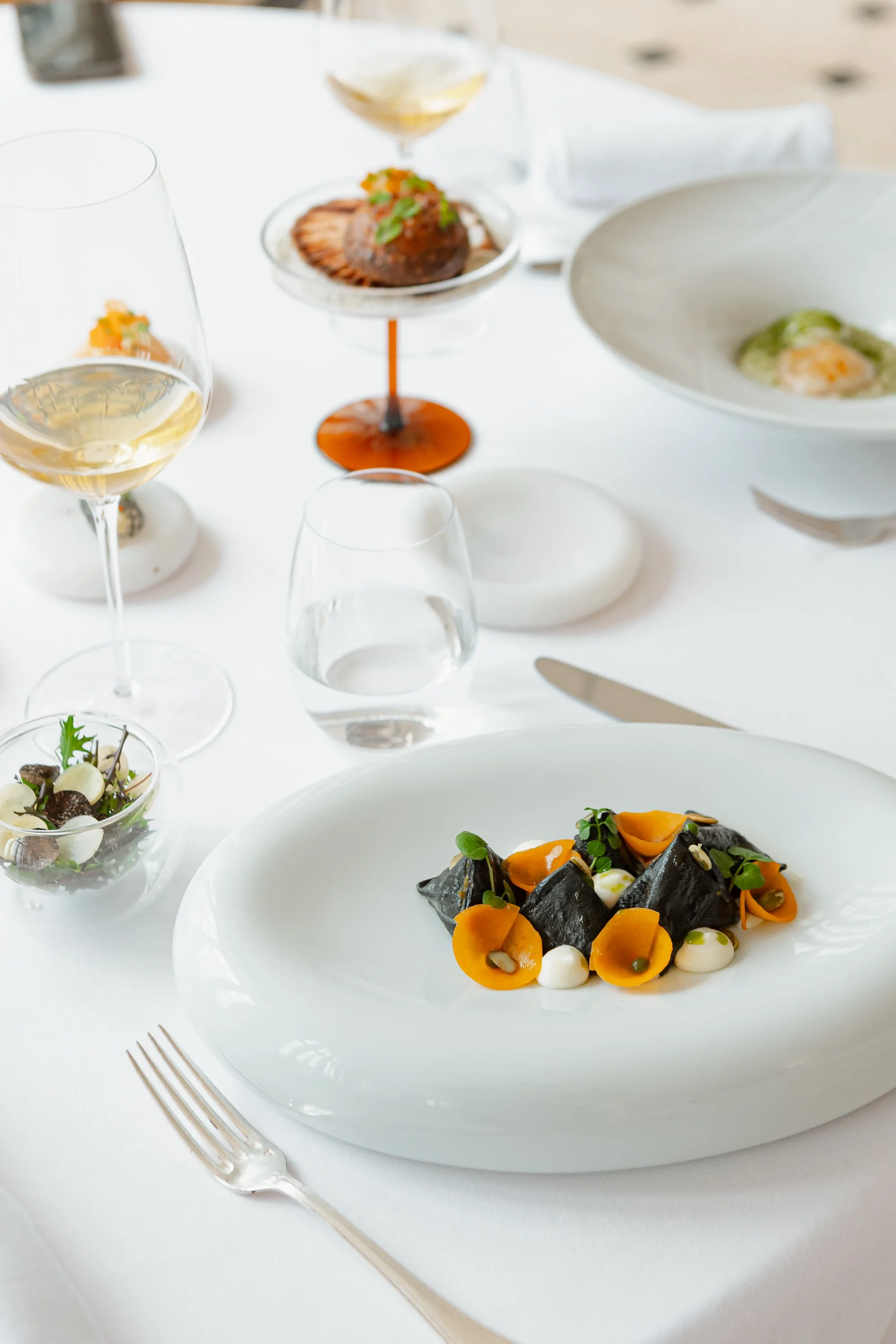 Une table élégante avec un plat principal de pâtes noires avec des feuilles vertes, des fleurs orange et de petites sauces blanches, accompagnée de plusieurs verres de vin blanc et d'autres plats sophistiqués.