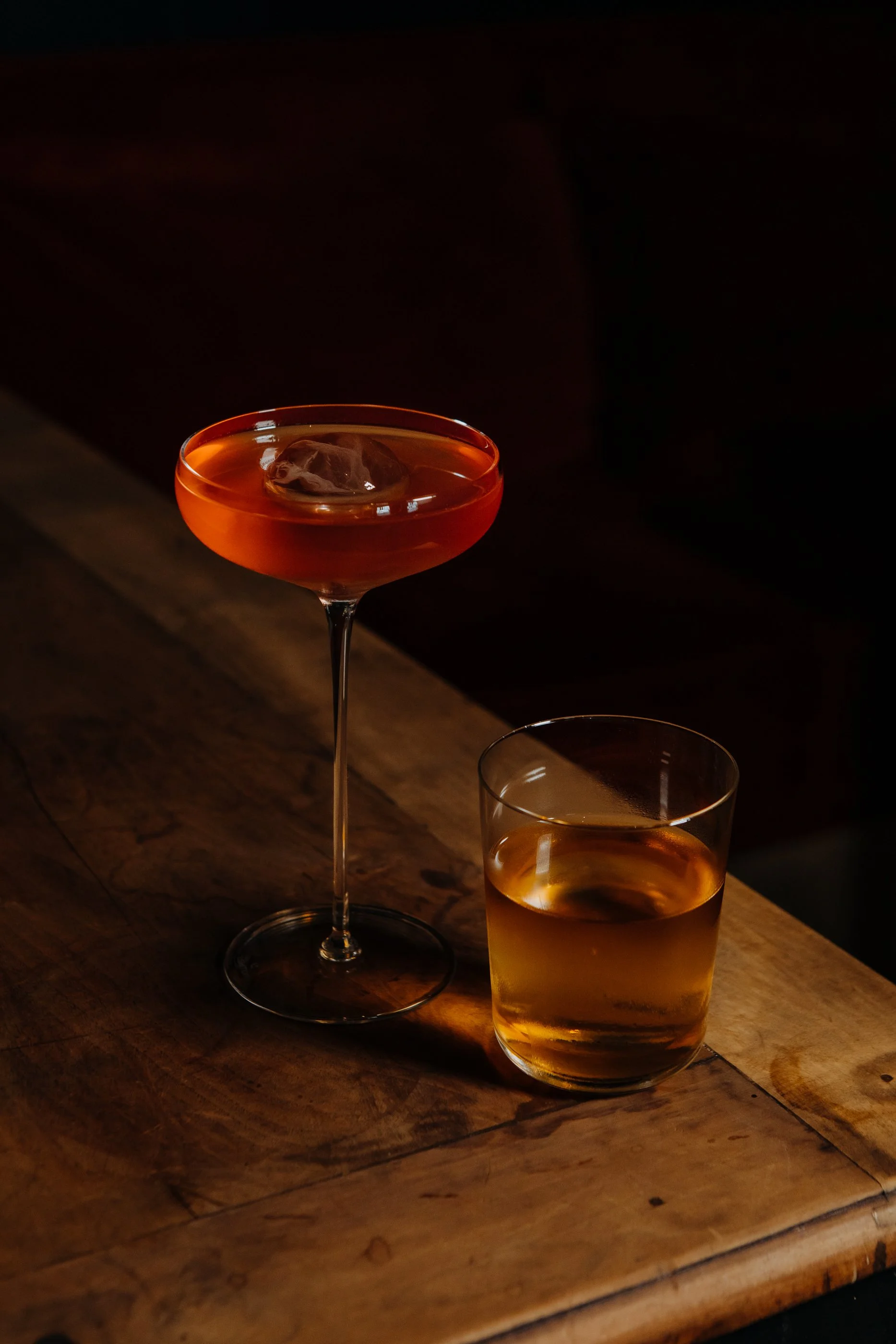 Deux cocktails, l'un dans un verre à martini avec une glace, l'autre dans un verre tumbler, posés sur un plan en bois dans un environnement sombre.