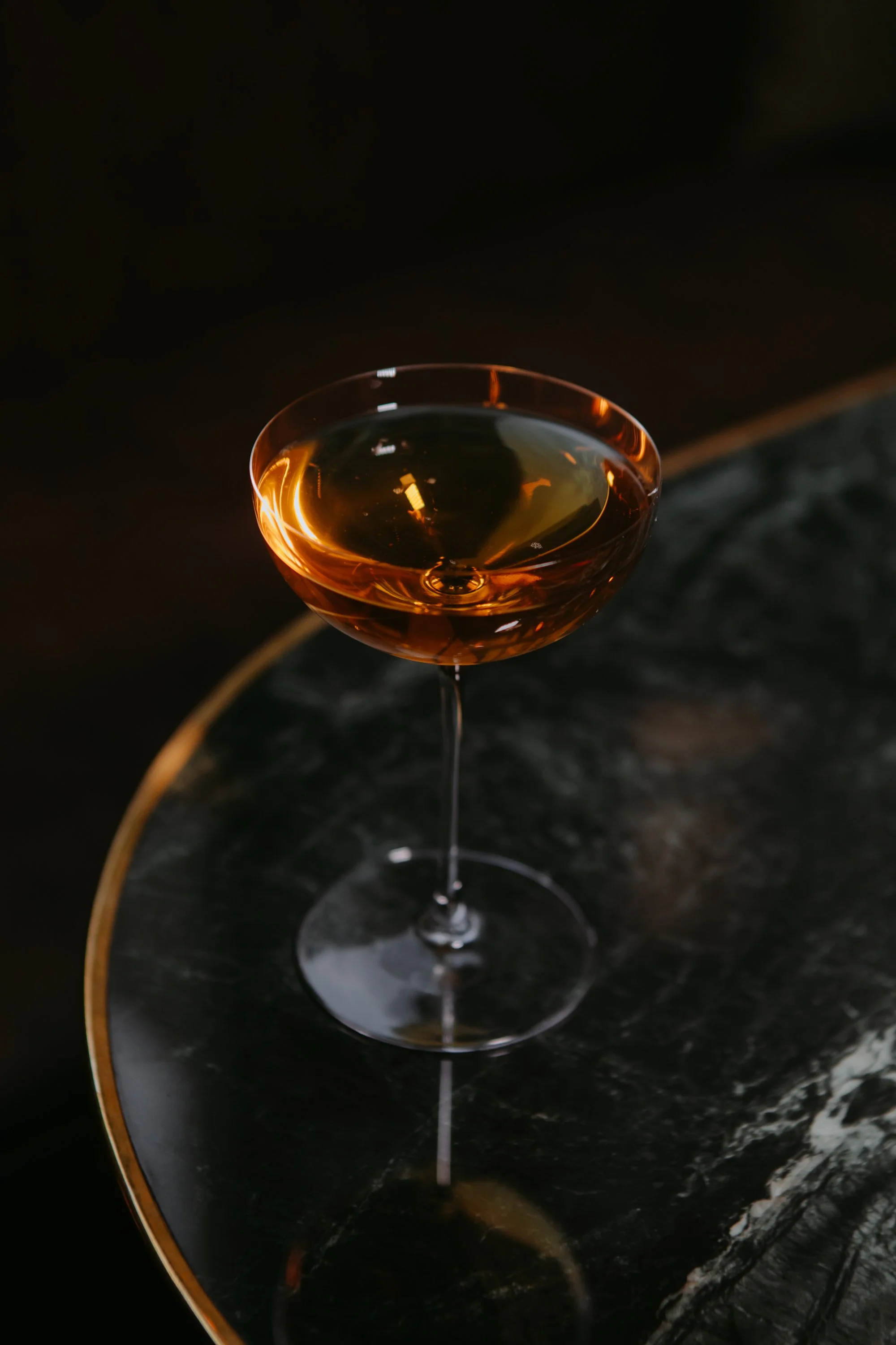 Un verre de cocktail rougeâtre placé sur une surface noire brillante avec un fond sombre.
