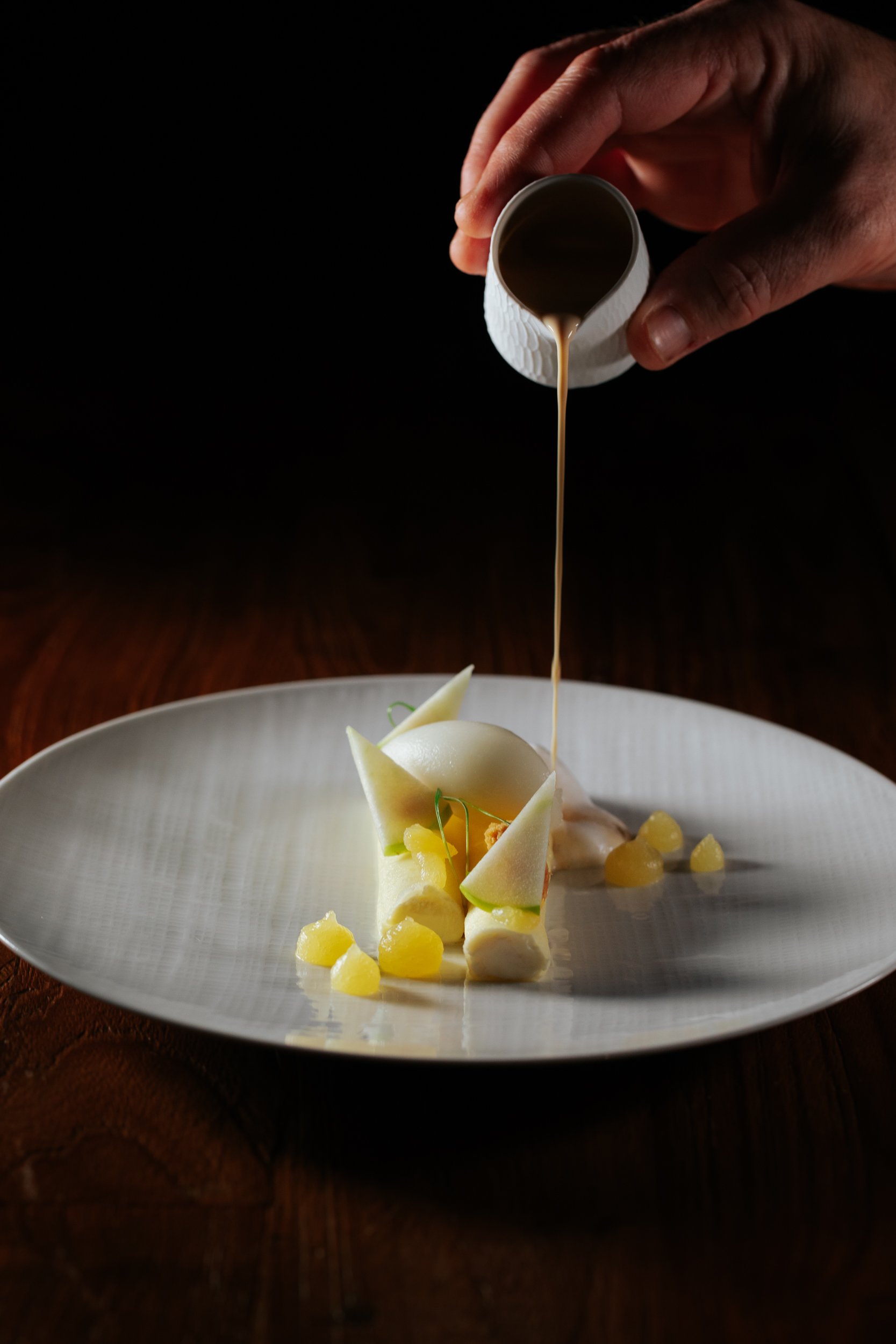 Une main verse une sauce brune sur un plat de dessert sophistiqué avec une boule de glace, des morceaux de fruit et des décorations en forme de triangles sur une assiette blanche.