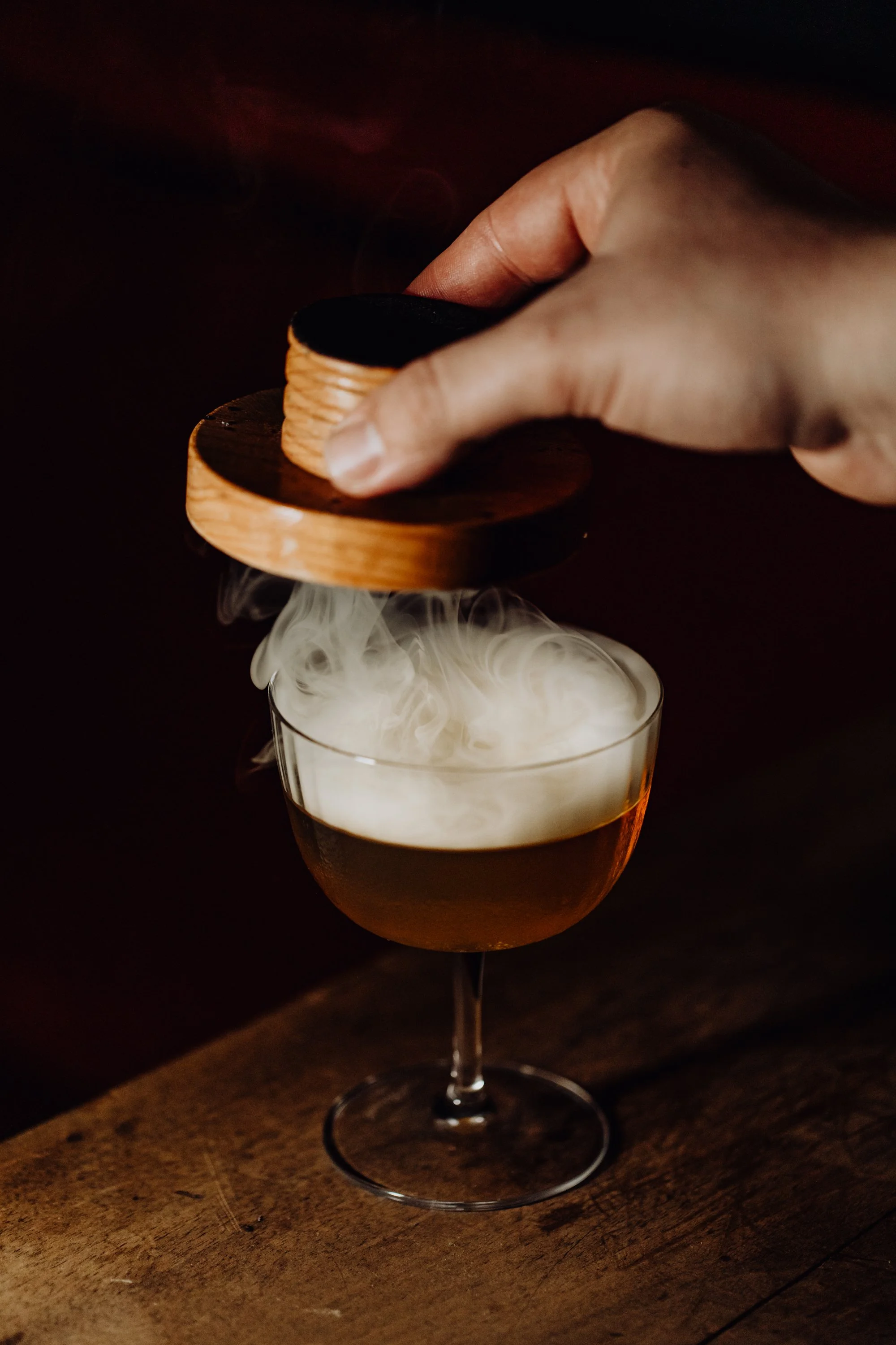 Person verse un verre de cocktail avec de la fumée s'échappant, recouvert par un plateau en bois