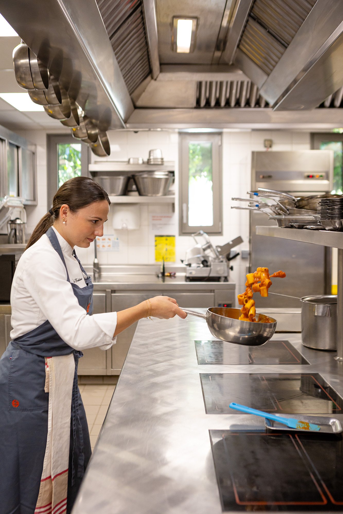 Une femme en uniforme de cuisine en train de faire sauter des pâtes dans une poêle en cuisine professionnelle.