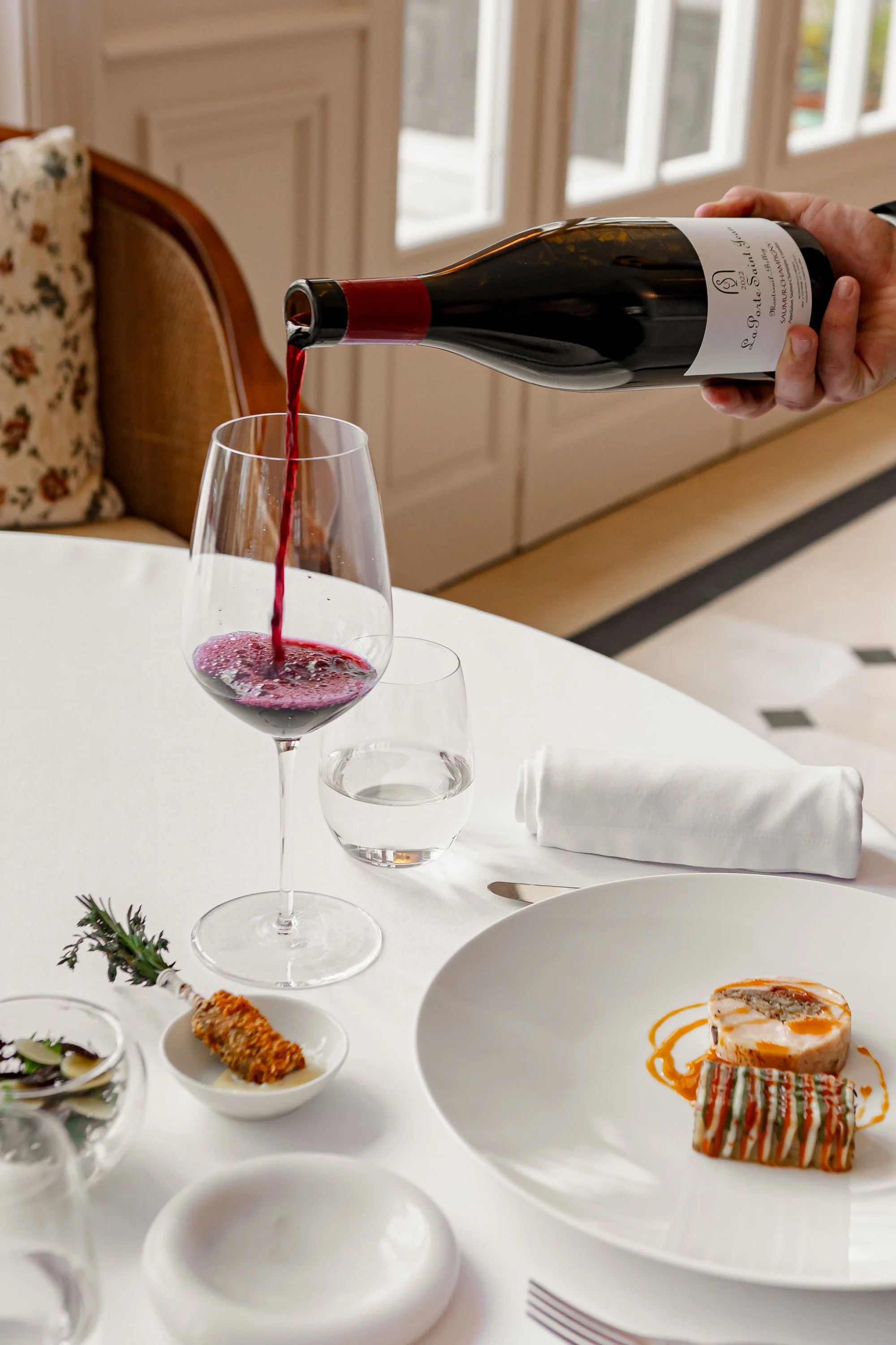 Une main verse du vin rouge dans un verre à vin sur une table de restaurant élégante, avec des plats, des verres d'eau, et une nappe blanche, dans un intérieur lumineux.