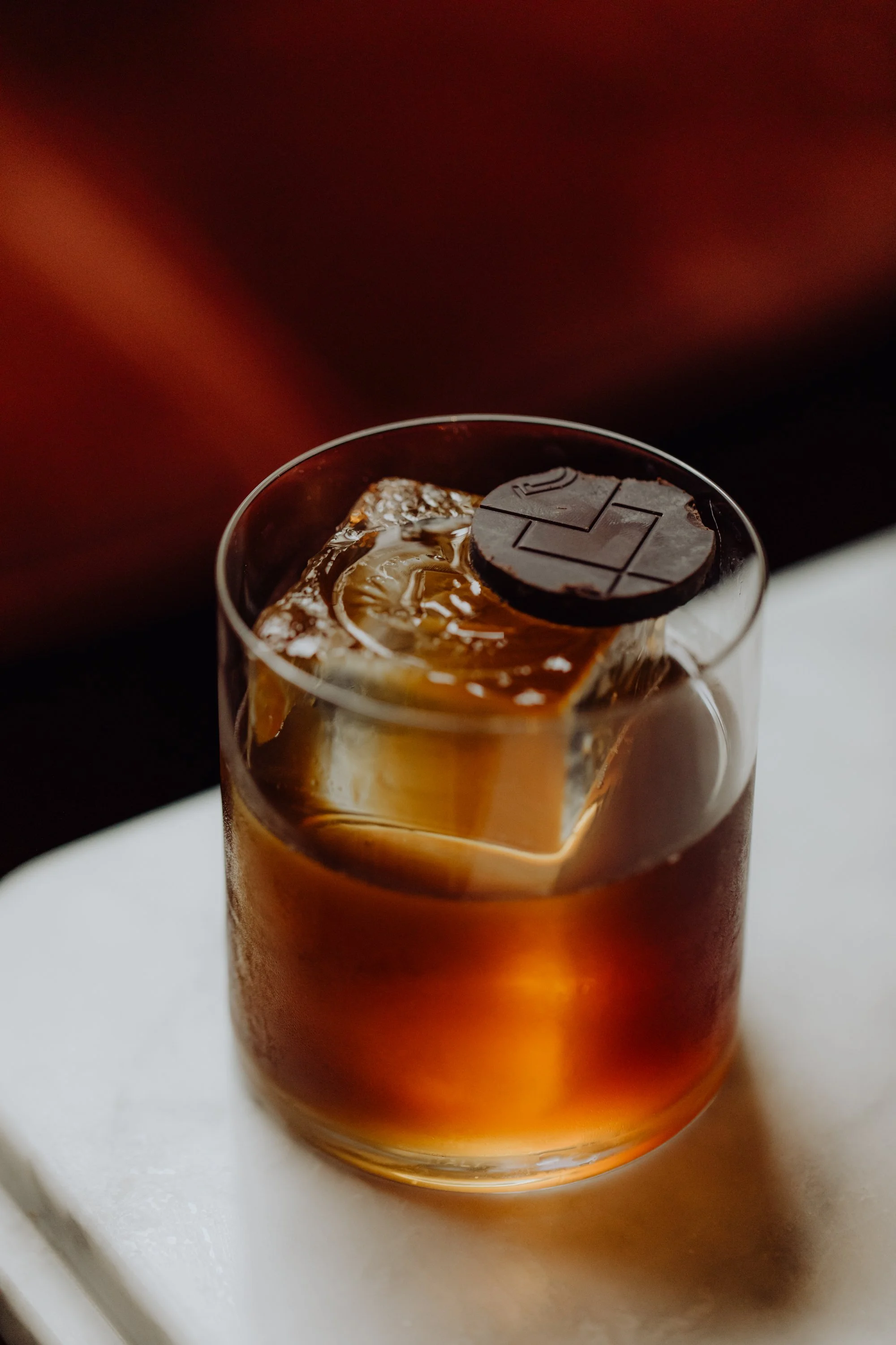 Verre de whisky avec un gros glaçon et un morceau de chocolat noir posé dessus.