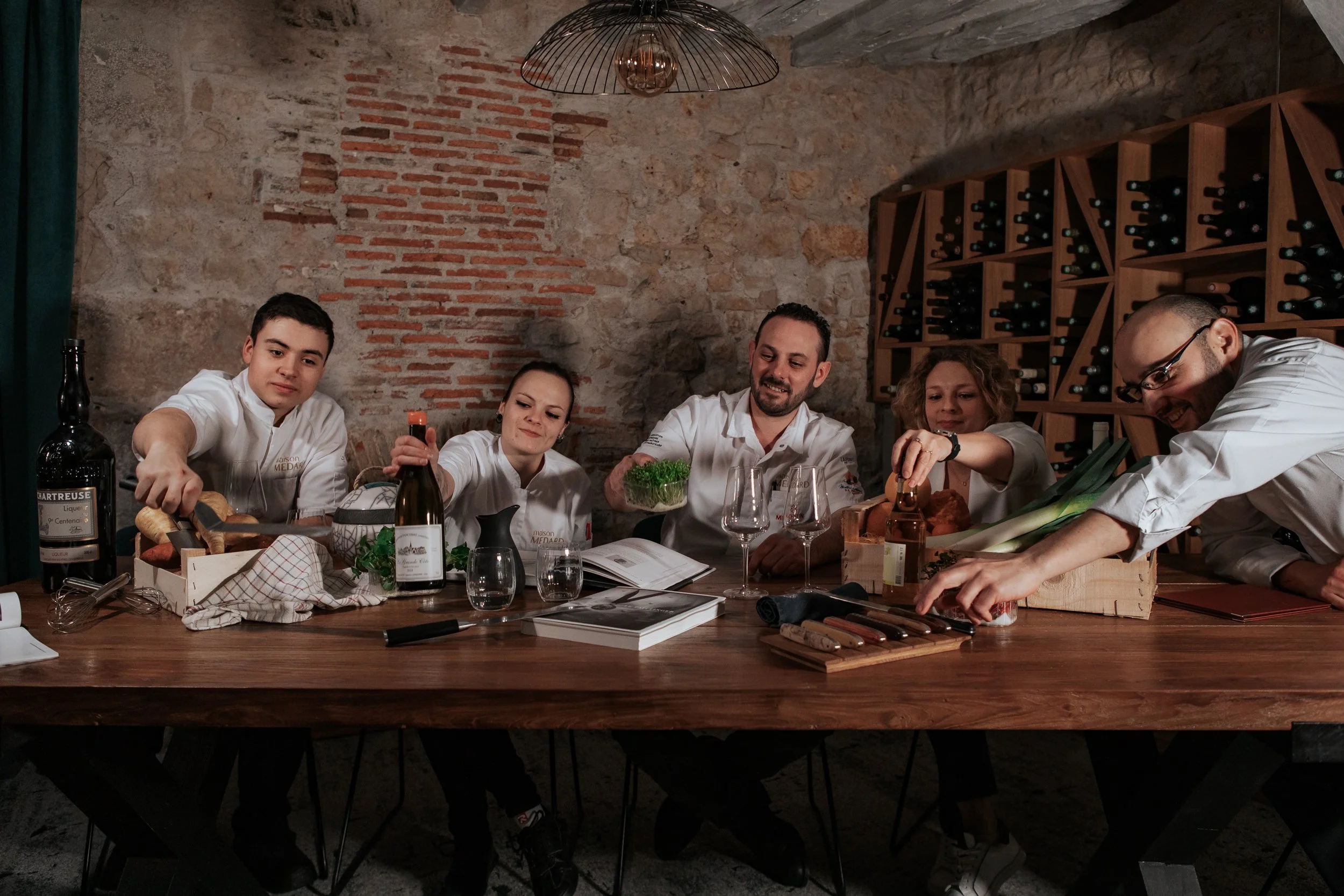 Groupe de chefs cuisiniers en team-building, travaillant autour d'une grande table en bois dans une cave à vin avec des murs en pierre et des étagères remplies de bouteilles de vin.