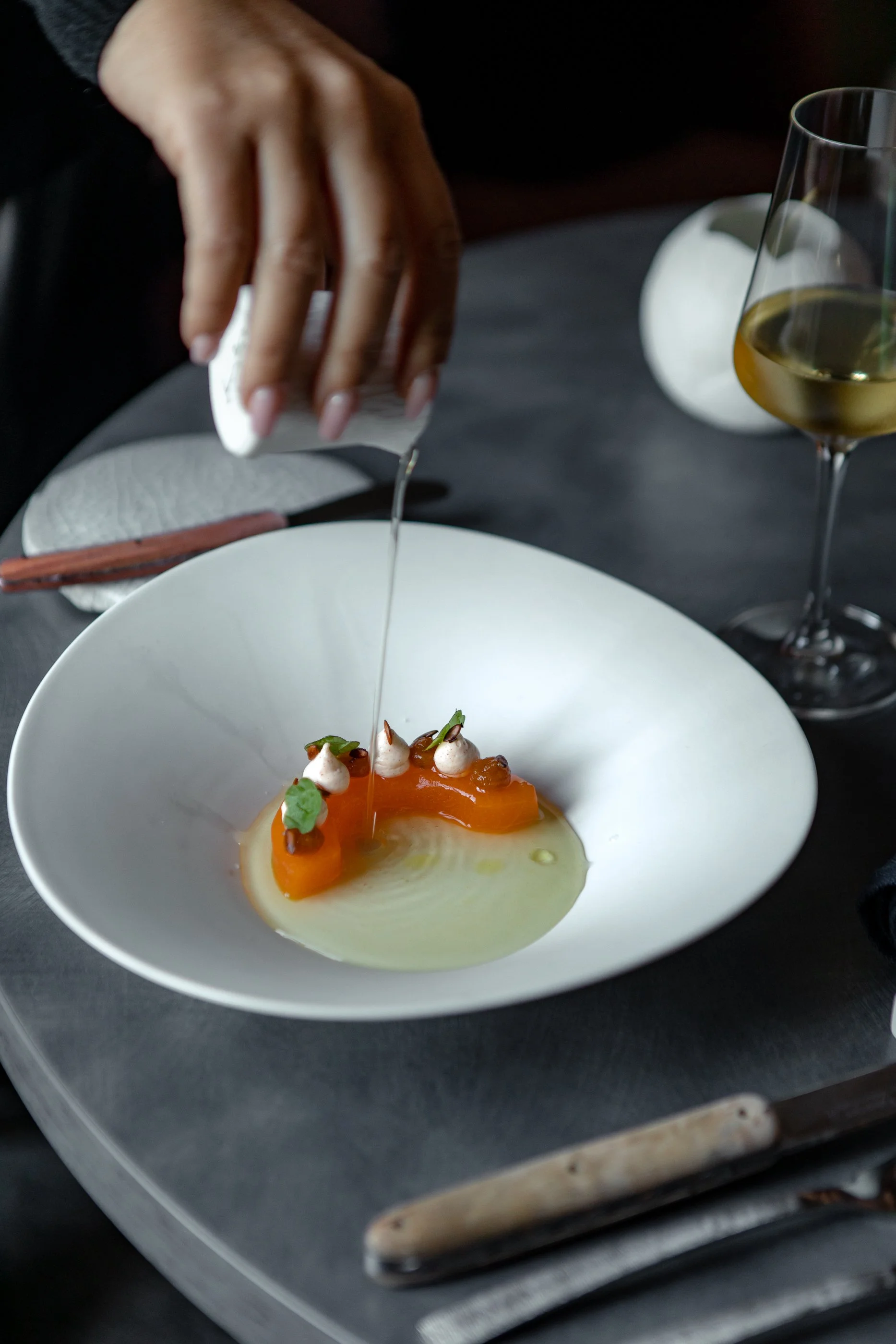 Une main verse de l'huile sur un plat sophistiqué contenant des morceaux d'aspégus et des meringues, avec un verre de vin blanc à côté.