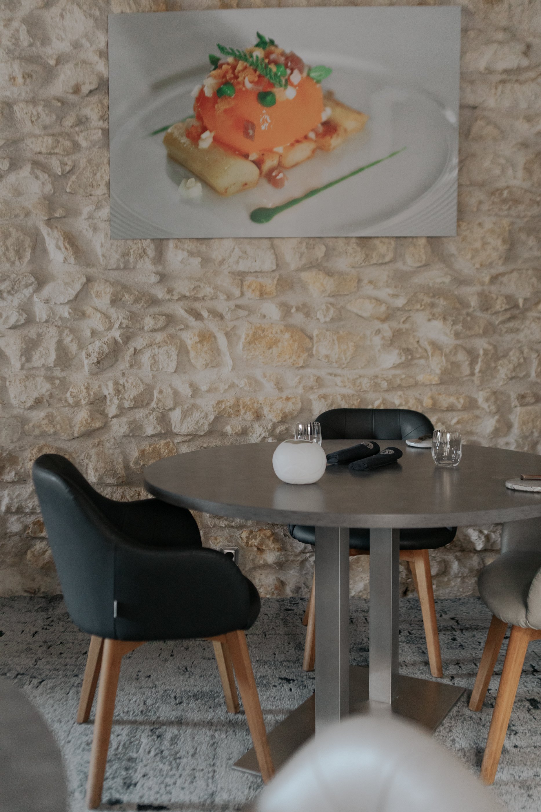 Une table ronde avec des chaises en cuir noir et beige, une carafe d'eau et des verres en verre sur la table, un mur en pierre en arrière-plan, et une photographie d'un plat sophistiqué de cuisine sur le mur.