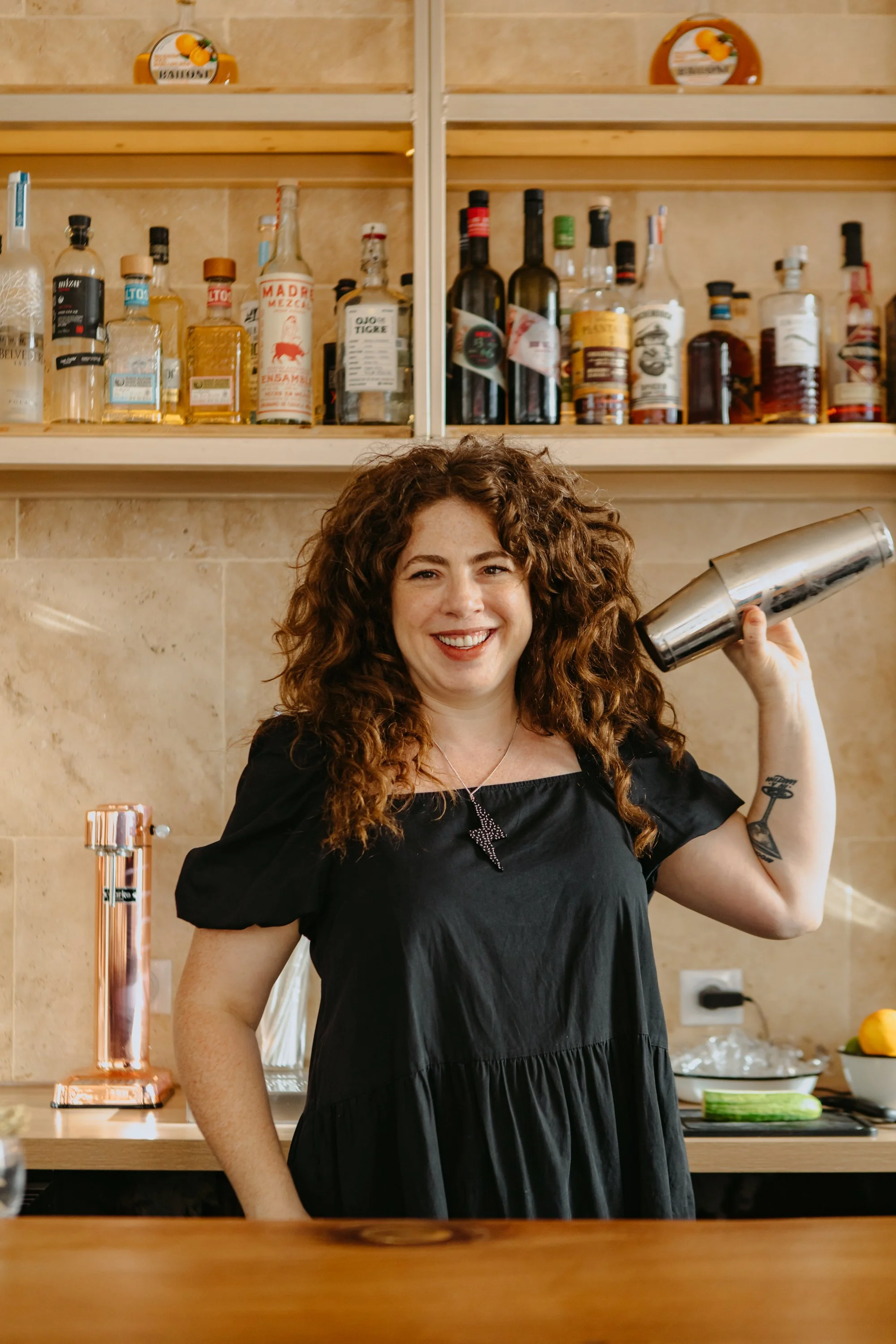 Une femme souriante avec des cheveux bouclés, portant une robe noire, tenant un shaker à cocktail en métal, se trouvant dans une cuisine avec une étagère de bouteilles d'alcool derrière elle.