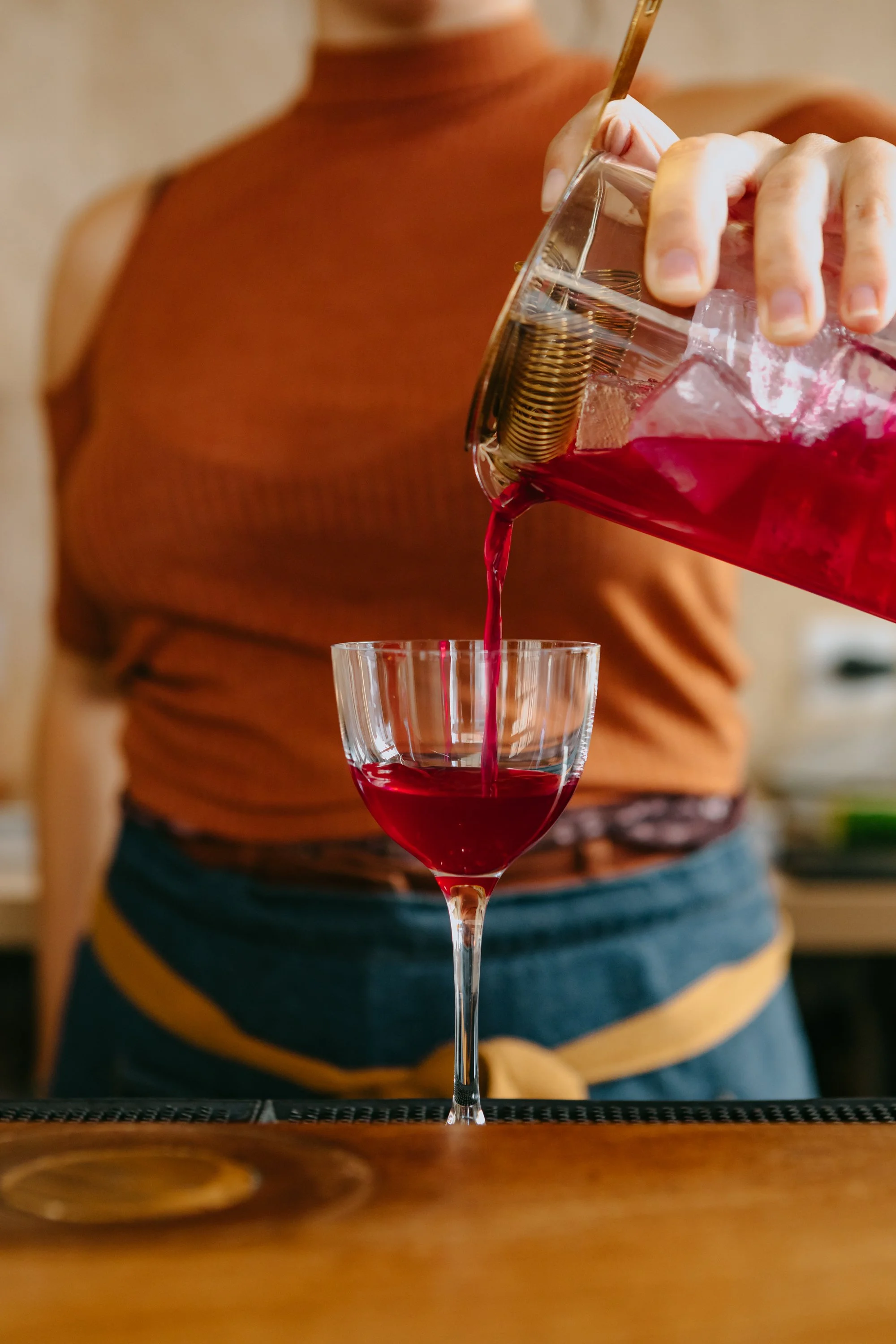 Une personne verse un cocktail rouge dans un verre à vin, la main tenant une carafe en verre avec un couvercle en métal.