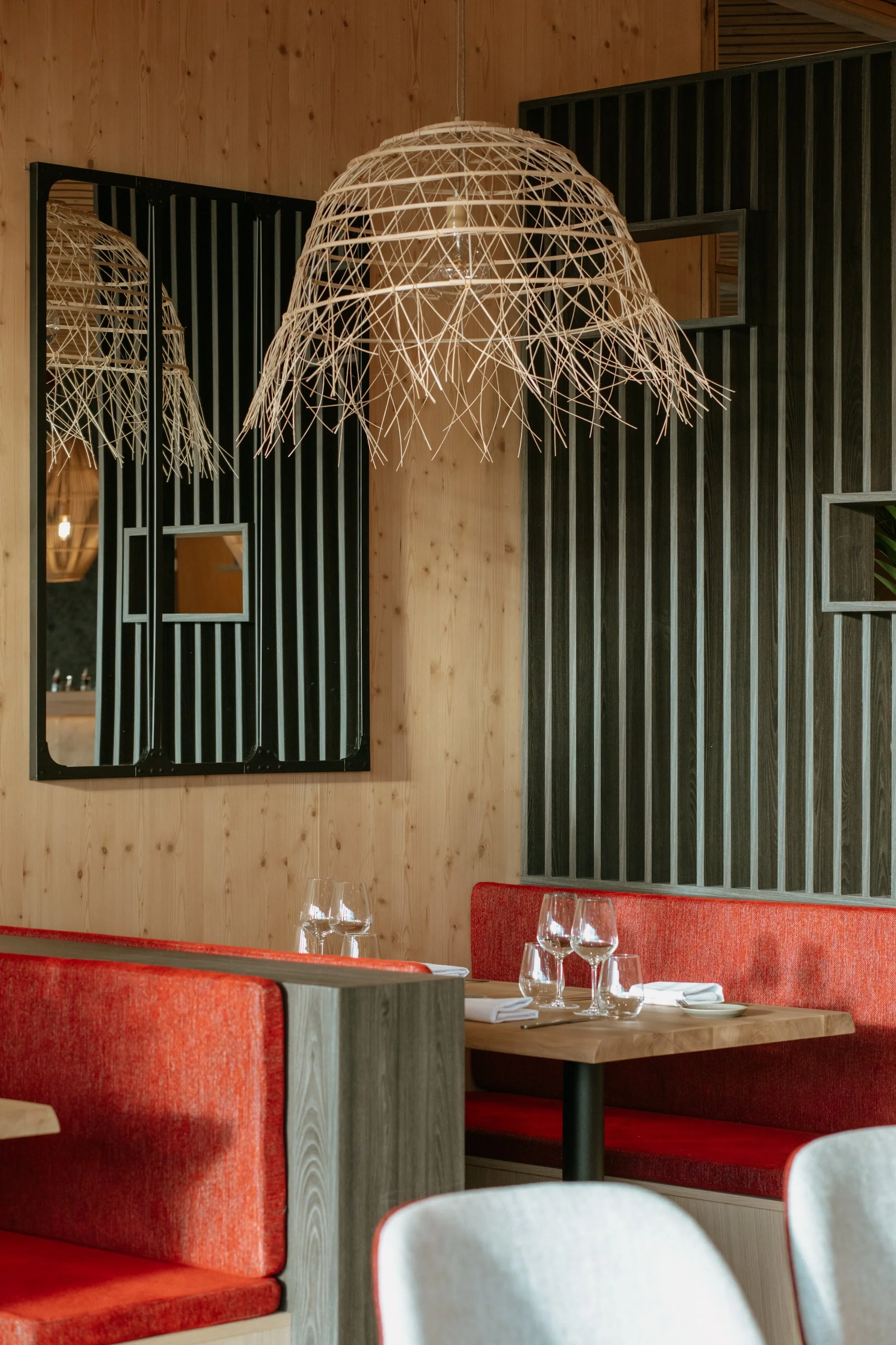 Intérieur d'un restaurant chic avec sièges en bois, banquette rouge, verres à vin et assiettes sur une table, lampes suspendues en osier, miroirs modernes et mur en bois