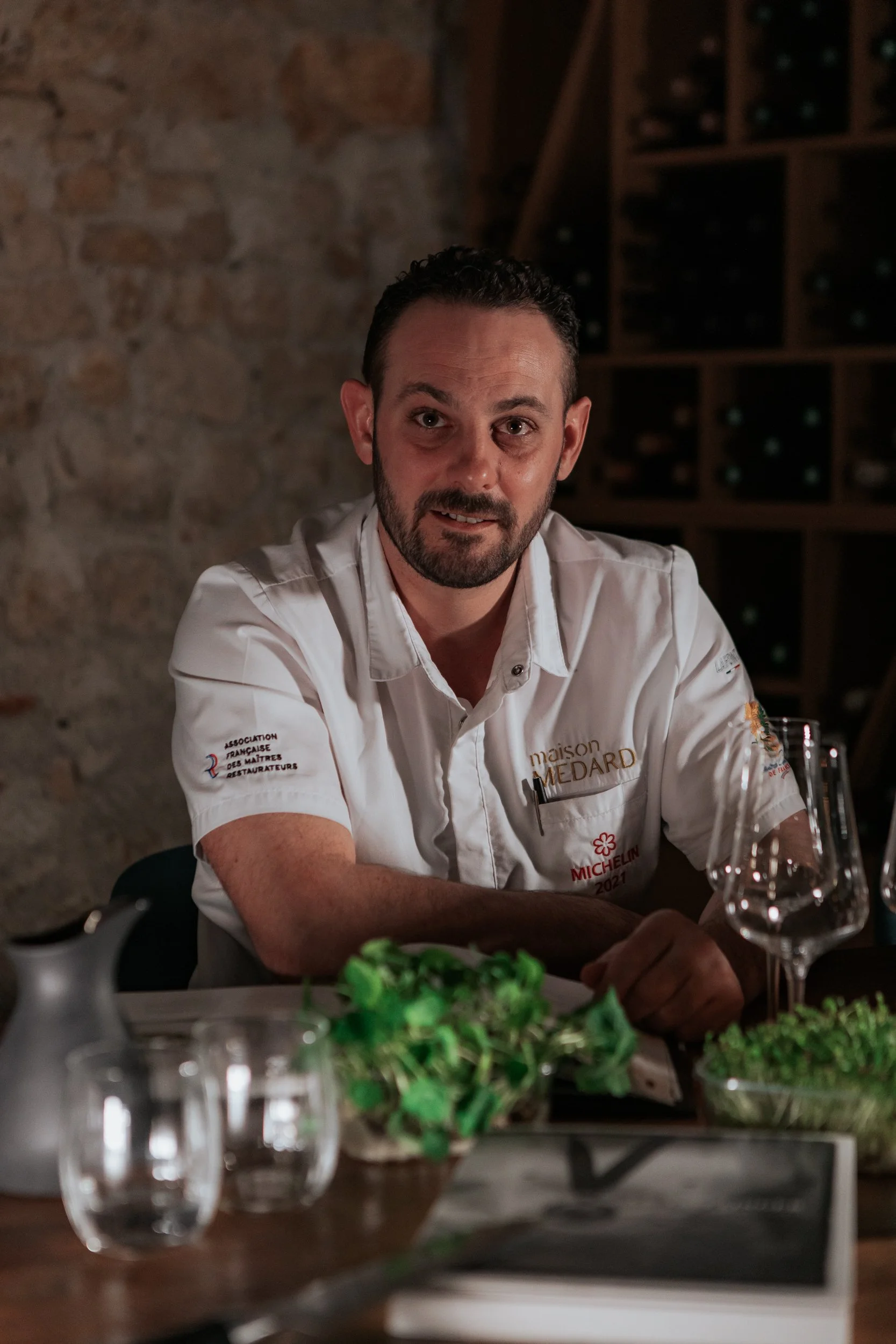 Un homme portant une chemise blanche avec des logos de la maison Medard et de Michelin, assis à une table dans un restaurant ou une cave à vin, entouré de verres et de plats, avec un mur en pierre en arrière-plan.
