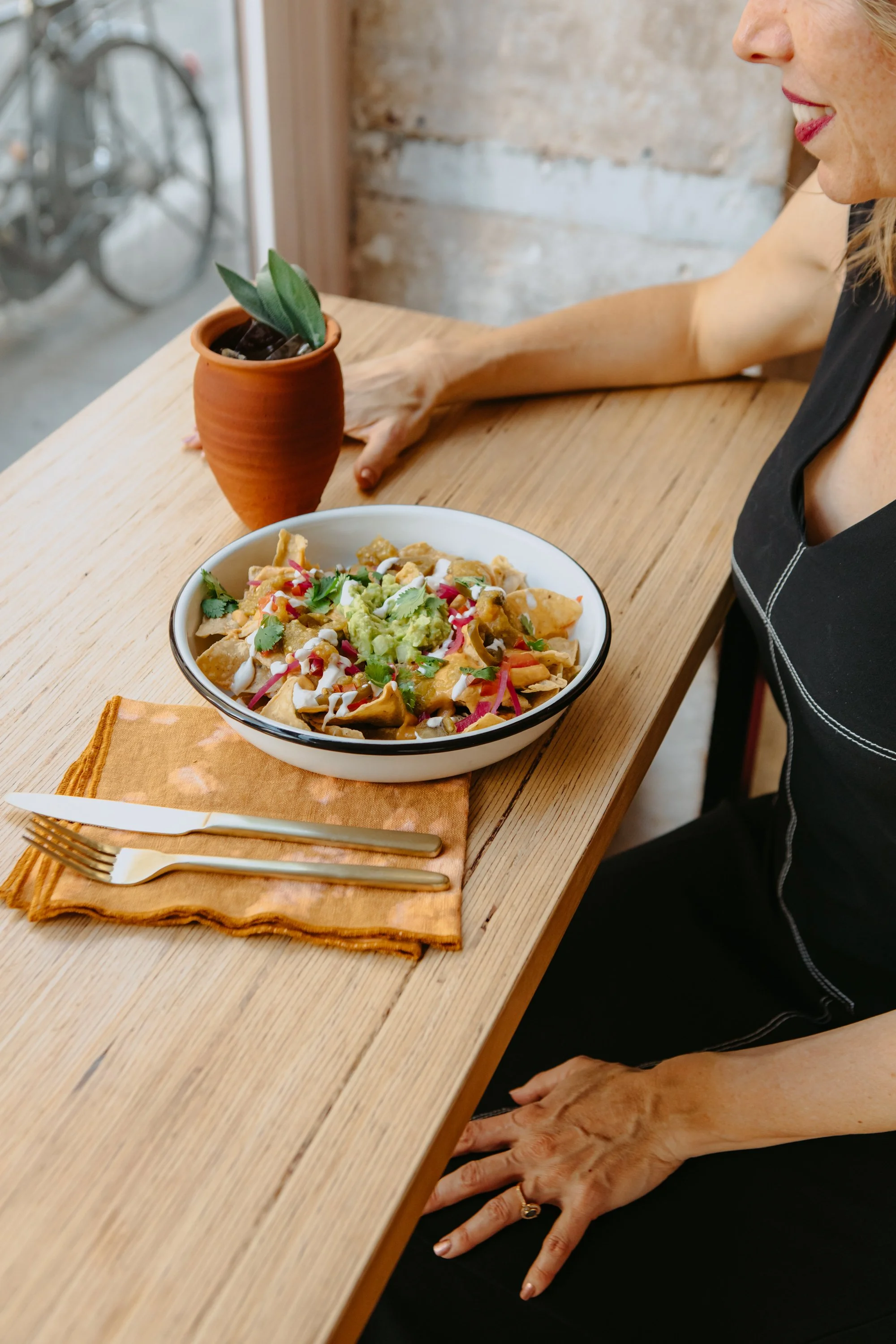 Une femme assise à une table en bois, devant un bol de nachos avec guacamole, crème, pico de gallo et coriandre, une nappe orange, une fourchette et un couteau en or, une plante en pot terracotta à côté de la tasse, un mur en briques en arrière-plan.