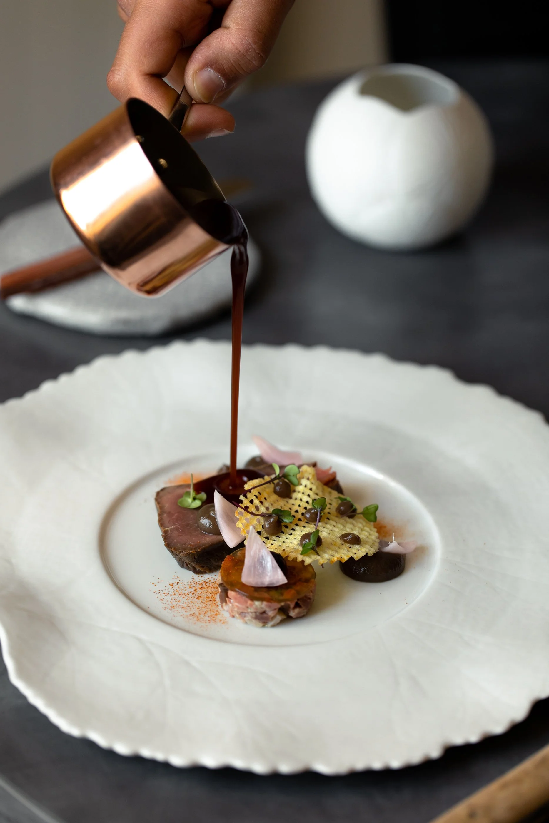 Une main verse une sauce chocolatée sur un plat de présentation gastronomique contenant plusieurs morceaux de viande, décorations délicates et croustillantes, ainsi que des petits éléments végétaux, sur une assiette blanche.