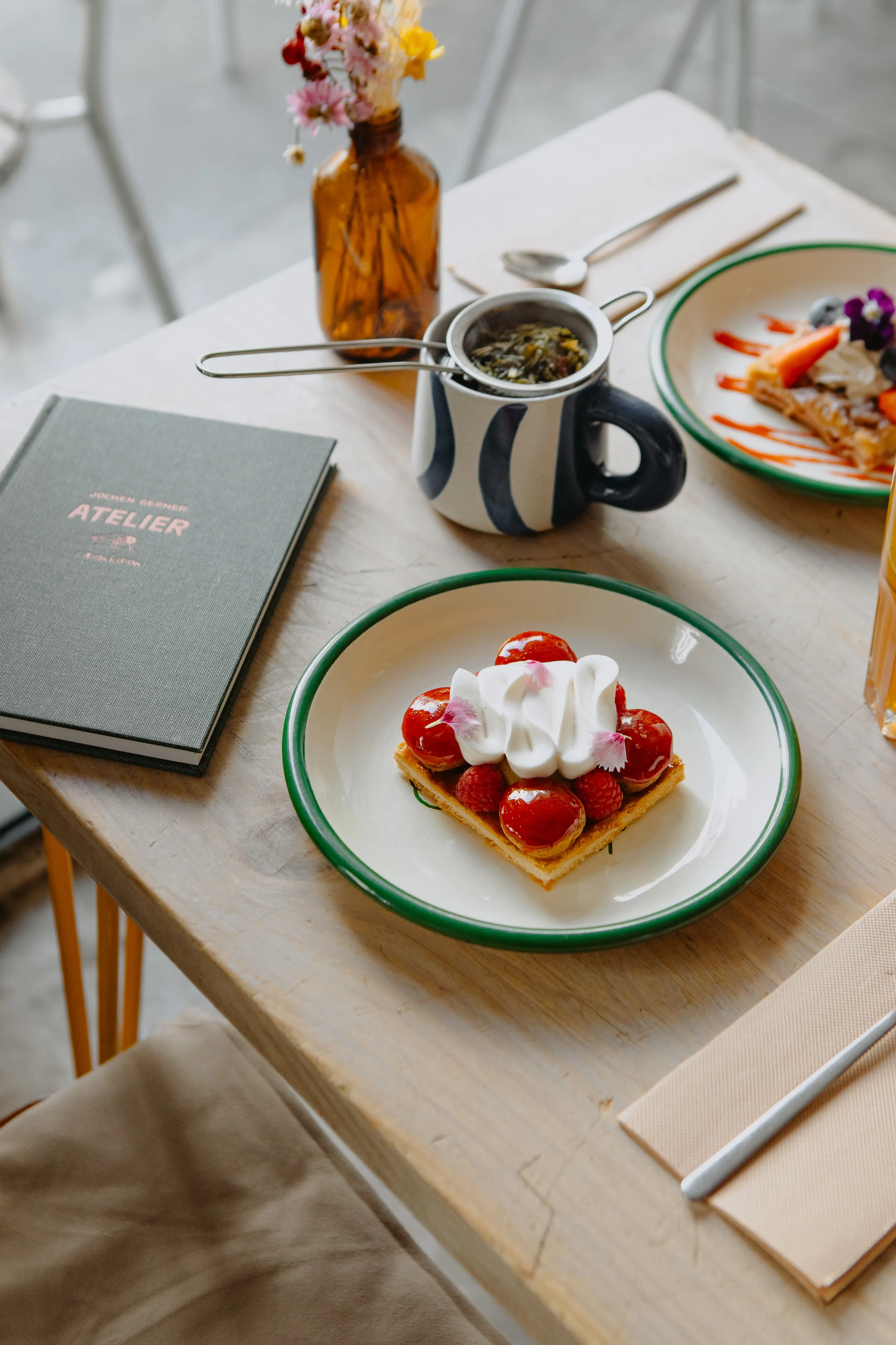 Une table avec une tarte aux fruits garnie de crème et de fruits rouges, une assiette avec un plat salé, une tasse avec une soupe, un carnet, une bouteille avec des fleurs, une cuillère et un couteau, et un verre d'orangeade.