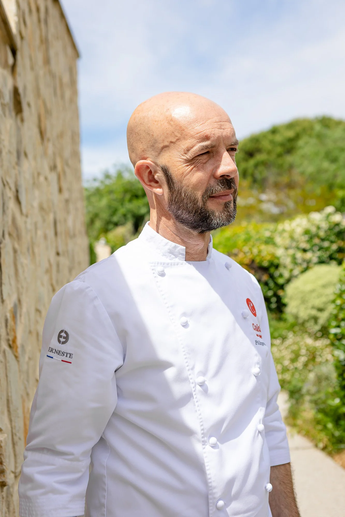 Un chef en uniforme blanc avec le nom Cole, se tenant à côté d'un mur en pierre, avec un jardin en arrière-plan.