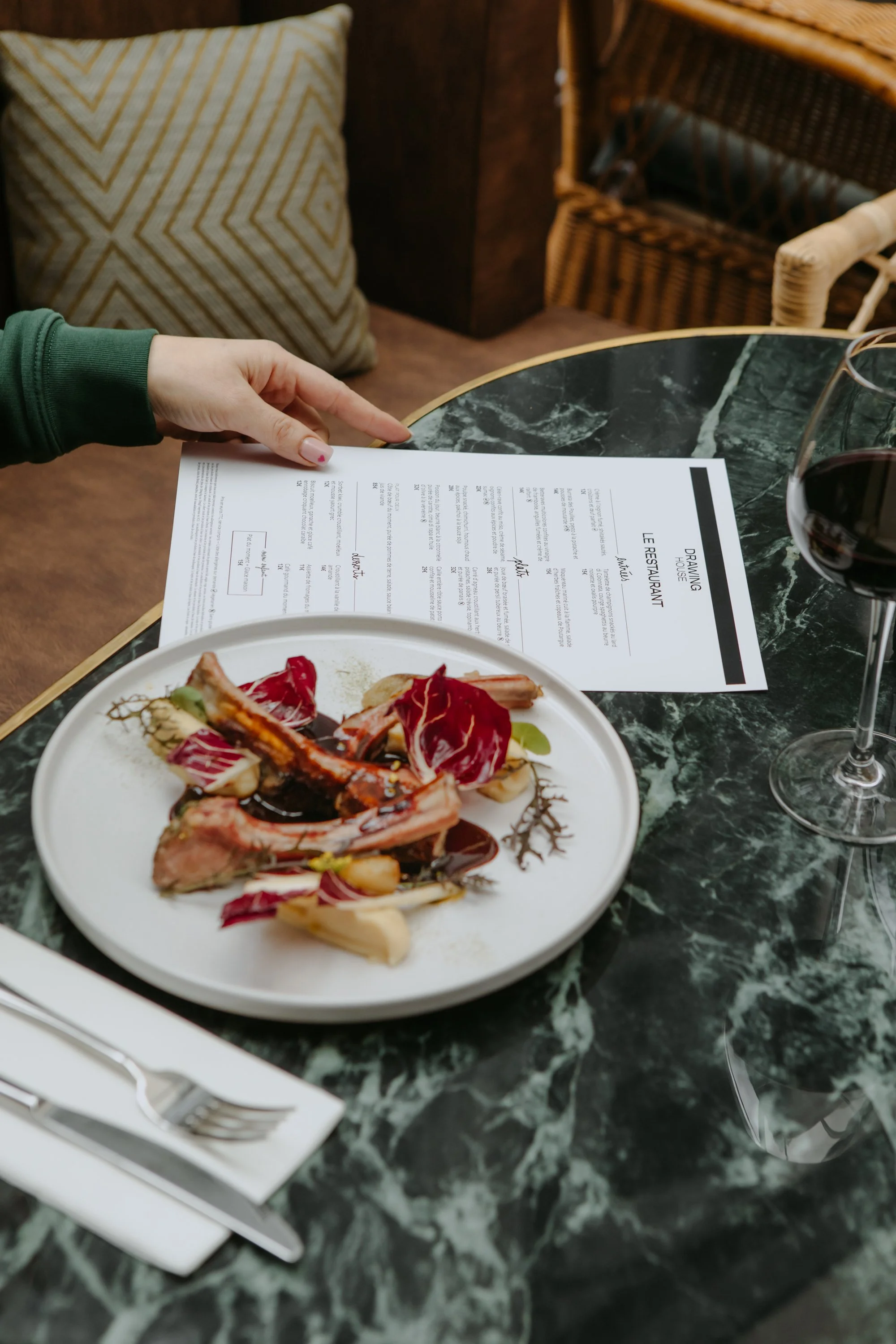 Assiette de nourriture avec des morceaux de viande, des légumes et une sauce, un verre de vin rouge, une femme regardant un menu dans un restaurant chic.