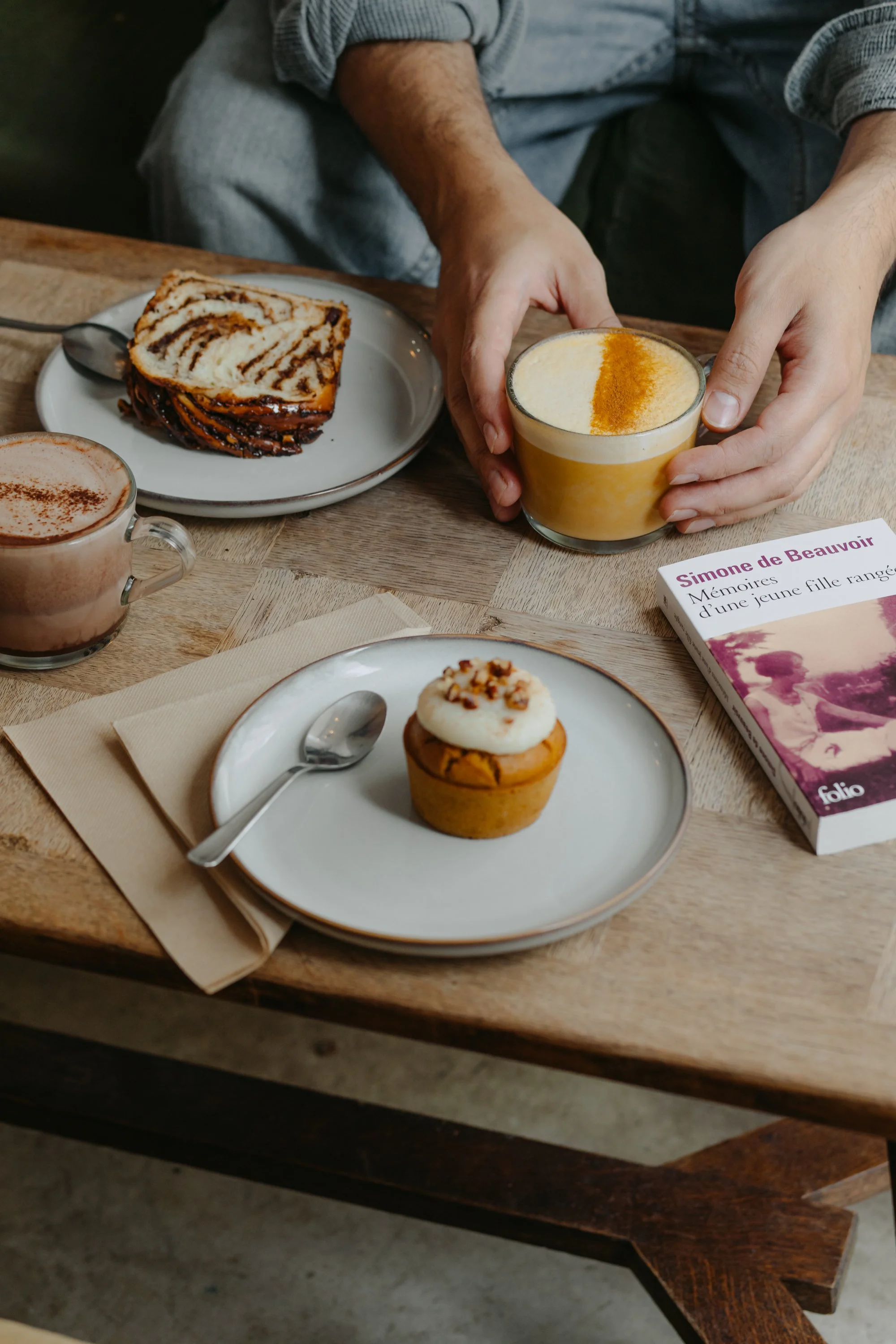 Gâteau roulé aux saveurs chocolat et vanille, cupcake à la vanille avec glaçage blanc et noix, tasse de chocolat chaud, verre de latte au curcuma, livre de mémoire de Simone de Beauvoir sur une table en bois.