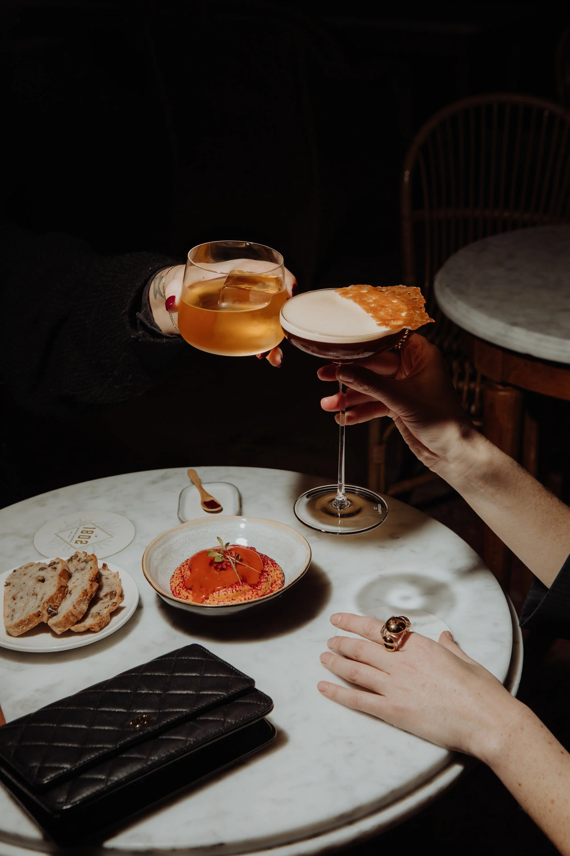 Deux personnes trinquent avec un verre de vin blanc et une coupe de cocktail, sur une table ronde en marbre avec des amuse-bouches, comme des tranches de pain, un dessert à la fraise, et un portefeuille en cuir