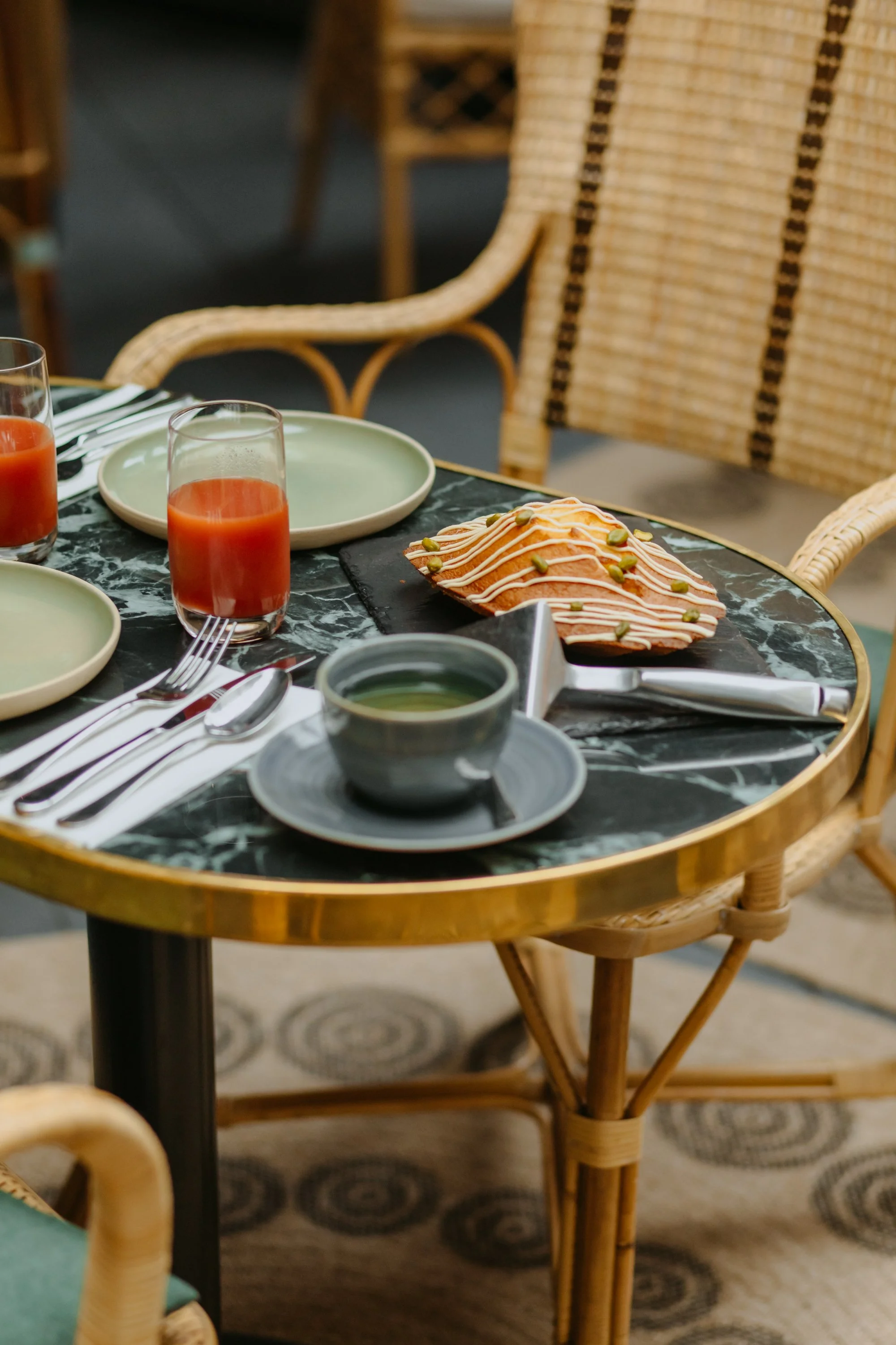 Une table ronde avec des assiettes, des verres de jus de tomate, une tasse de soupe et un plat de pâtisserie décorée de glaçage blanc et de graines de sésame, dans un environnement intérieur avec des chaises en rotin.