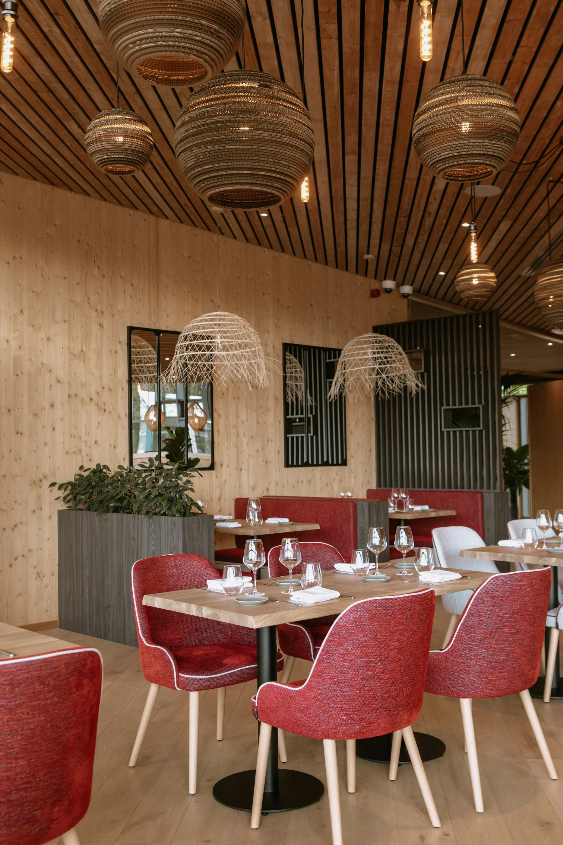 Salle à manger dans un restaurant avec des chaises rouges, des tables en bois, des verres à vin, de la vaisselle blanche, et un décor en bois avec des lampes suspendues