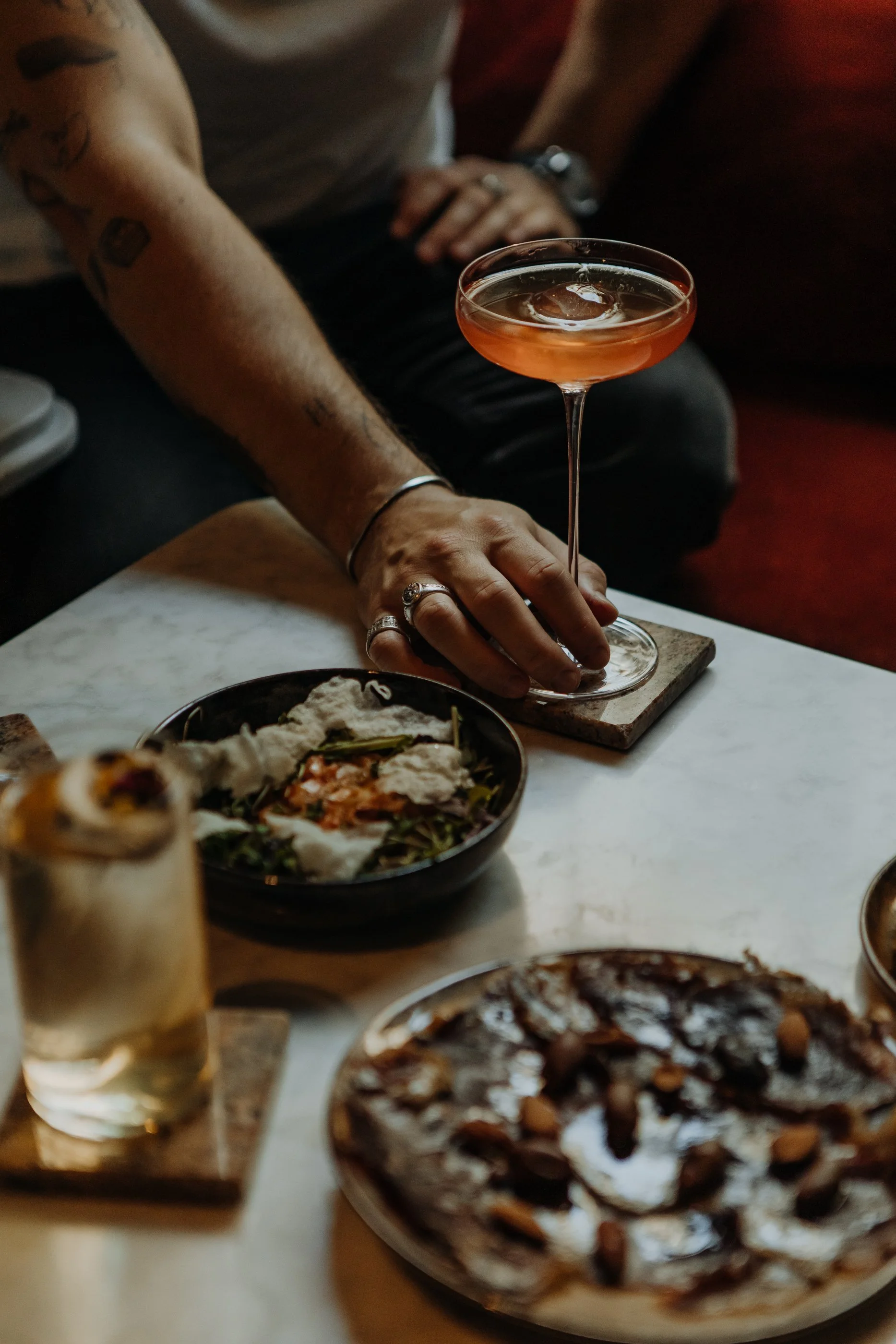 Une main tenant un verre à cocktail rose sur une table avec des plats de nourriture et des boissons.