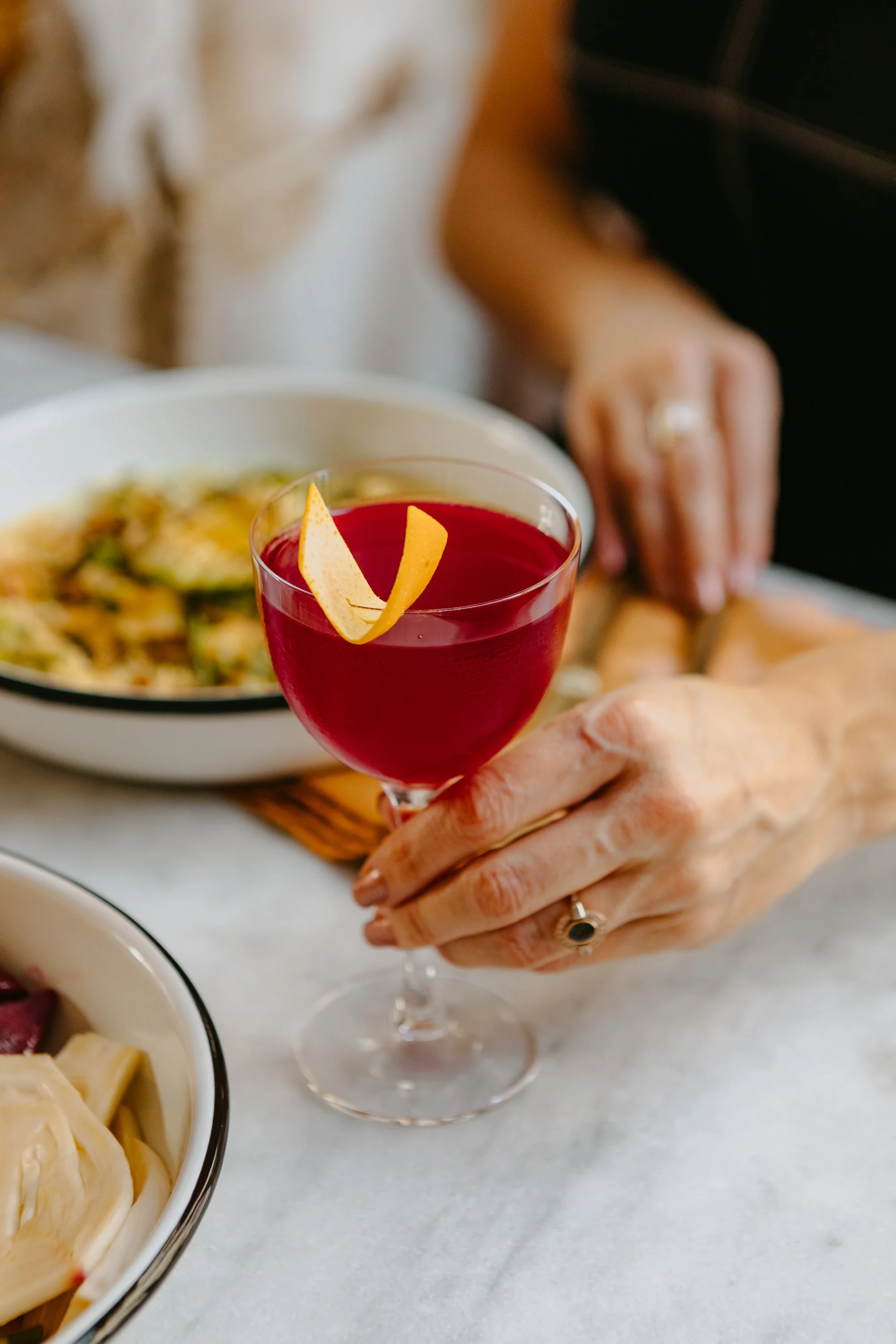 Une personne tient un verre de cocktail rouge avec une tranche de citron en garniture, avec un plat de nourriture flou en arrière-plan.