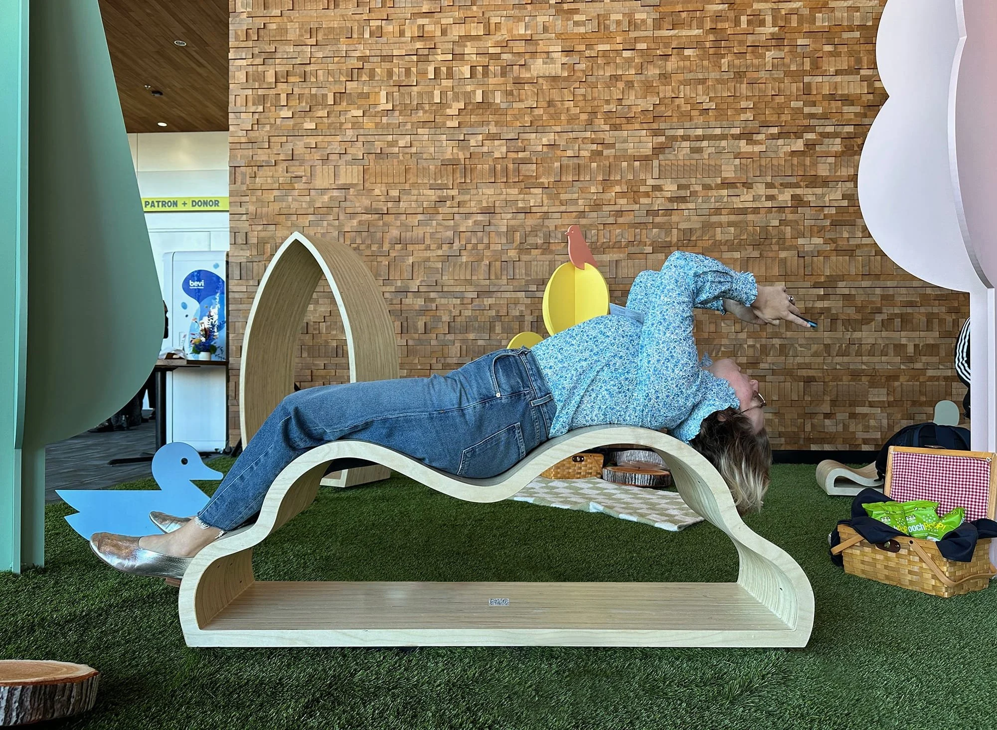 Person lying on a curved wooden lounge with a blue floral shirt and jeans, pointing to the right, on a green turf area with decorative wood and animal shapes in the background.