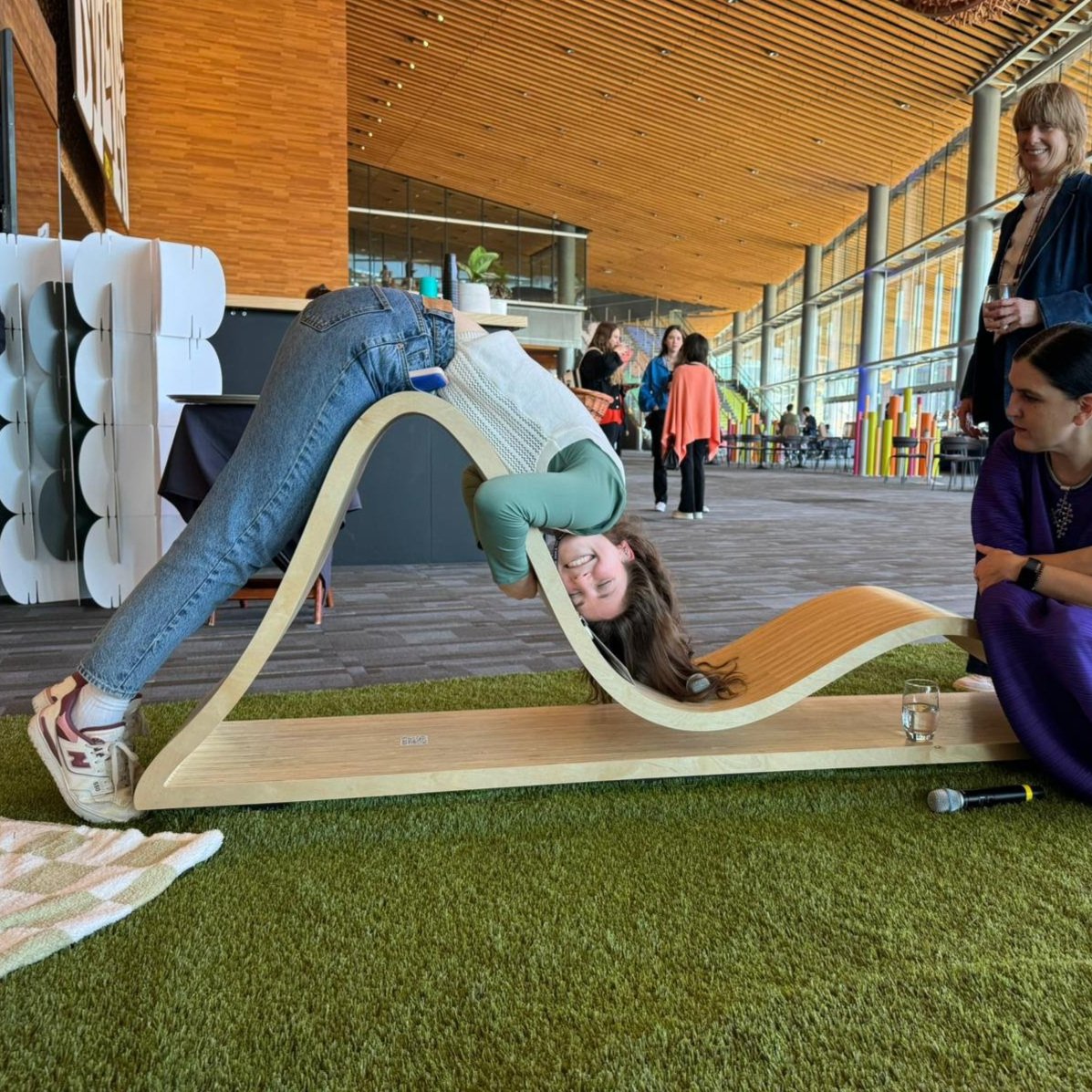 A woman is lying on a modern wave-shaped wooden bench in a public indoor space, with her head hanging upside down and smiling. She is wearing jeans and sneakers, and appears to be enjoying the moment. Several people are in the background, some standing and talking, others sitting, with a colorful and spacious environment featuring high ceilings and large windows.