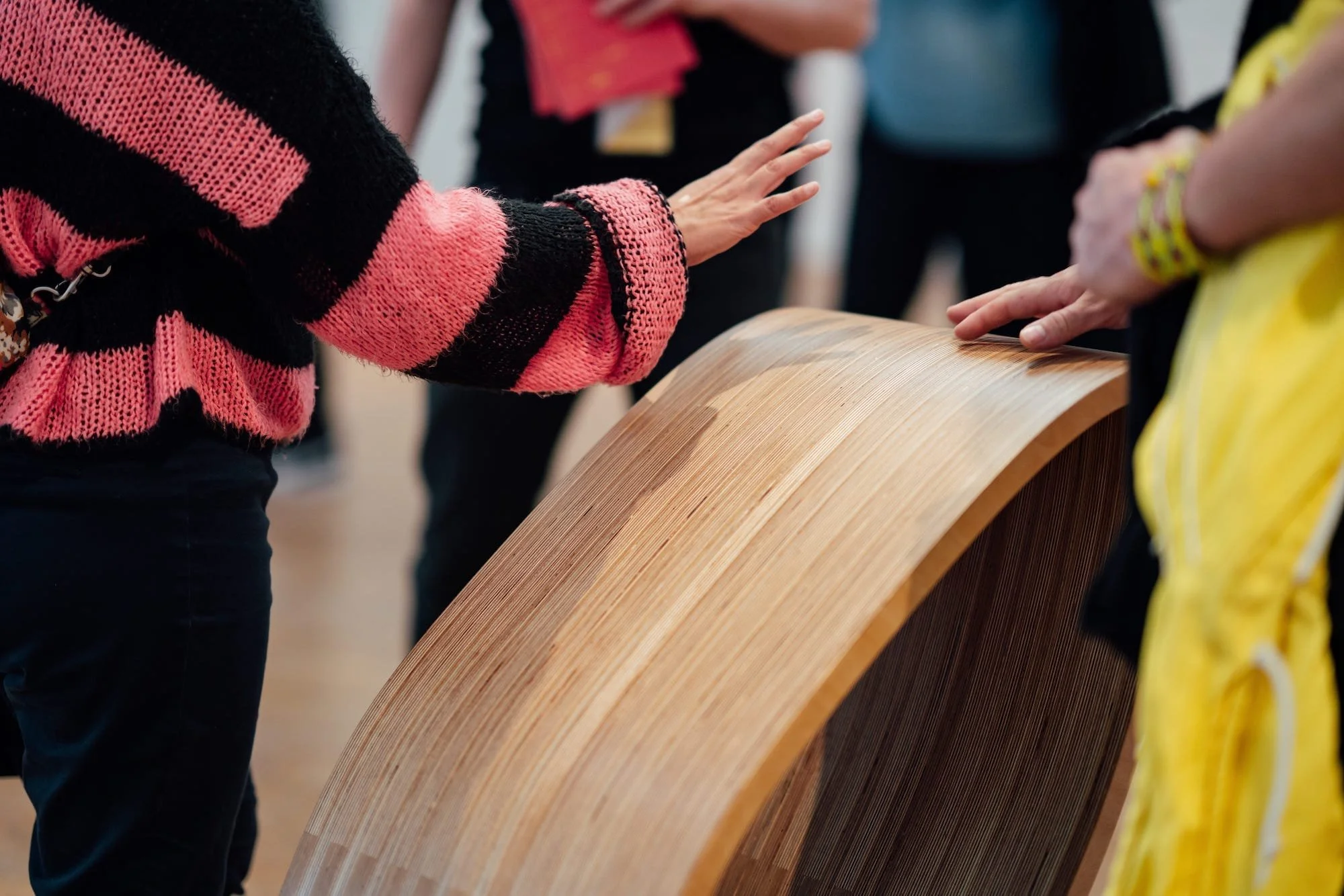 People touching a curved wooden sculpture or piece of furniture at an indoor event.