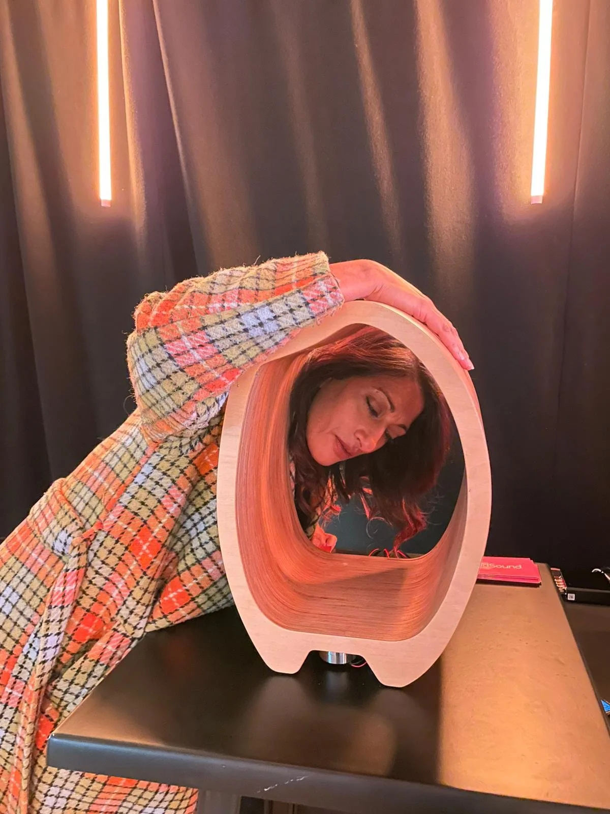 Woman leaning over a wooden speaker with a circular opening, with her face reflected in the wood, against a dark background with vertical light strips.