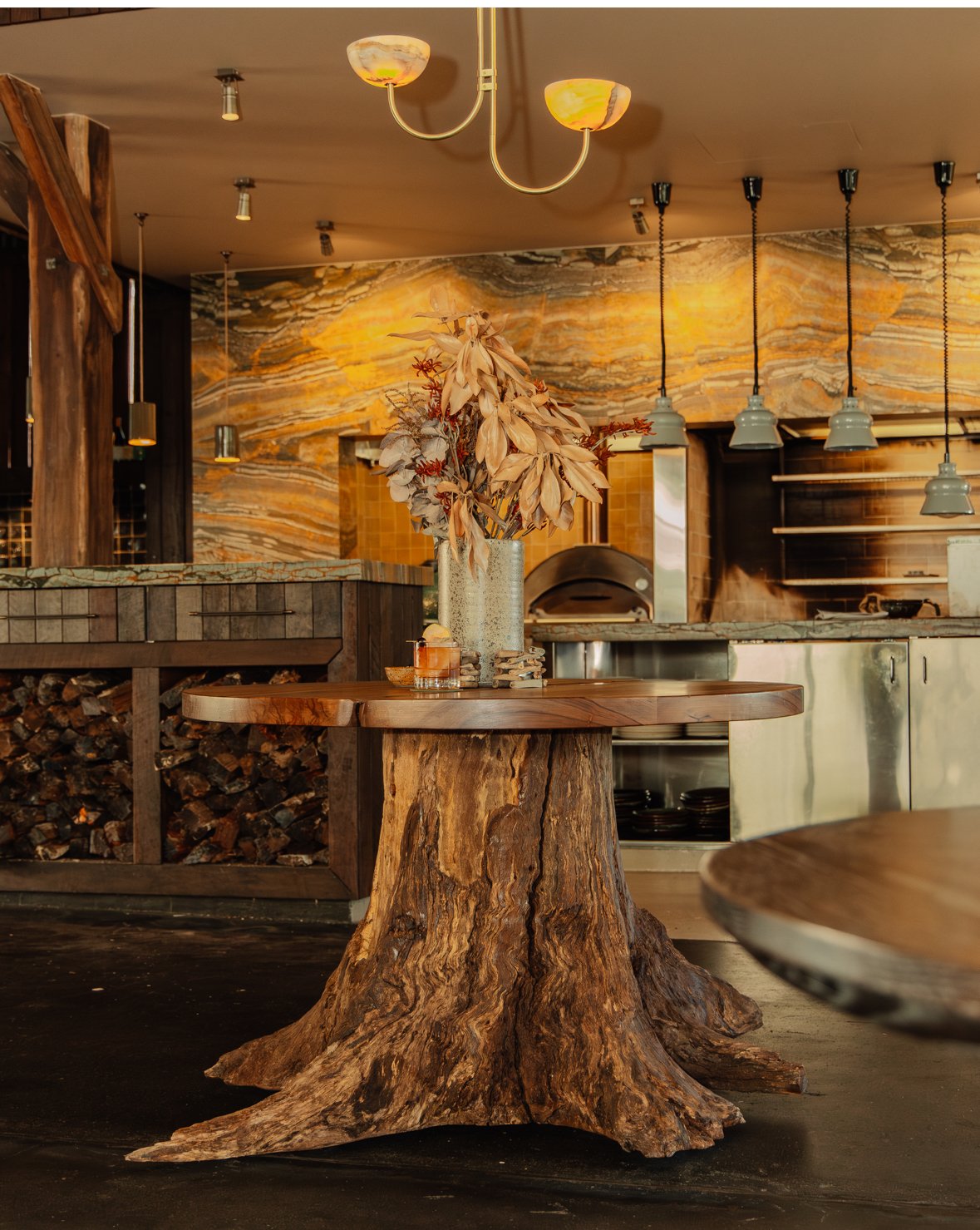 A large, weathered tree stump with roots visible, placed in front of a pile of cut tree logs and branches outdoors under a clear blue sky.