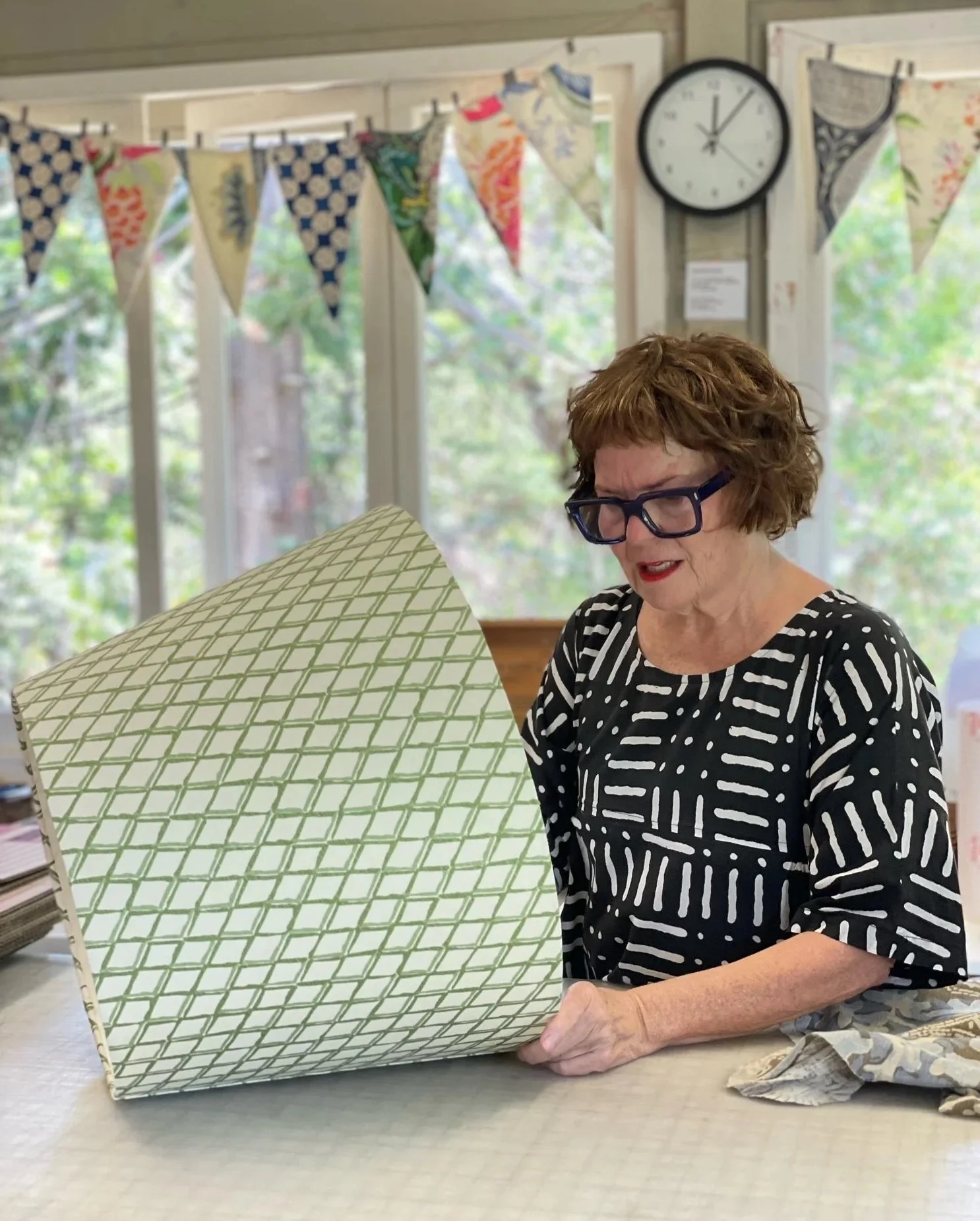 A woman with short curly hair wearing glasses and a black and white striped top is looking at a bespoke fabric lampshade with a green diamond pattern, sitting at a table in a bright room with windows, bunting.