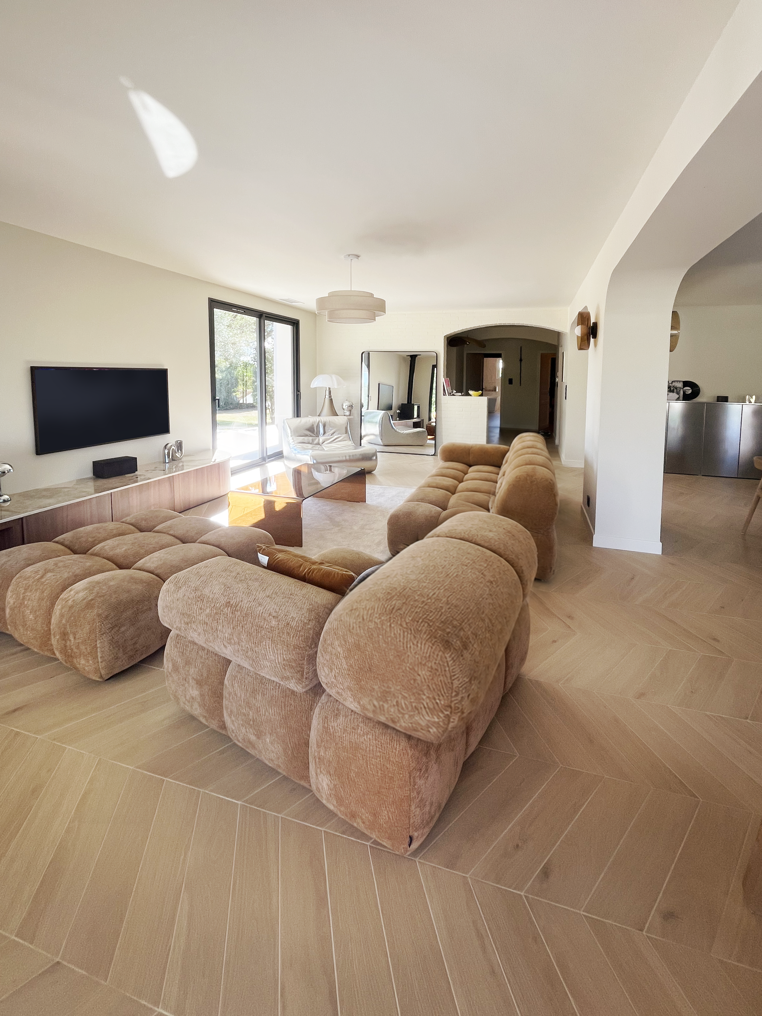 Un salon moderne avec un canapé en velours beige, une télévision à écran plat, une table basse en verre, une lampe de table, et un grand miroir. La pièce est lumineuse avec une porte-fenêtre donnant sur l'extérieur.