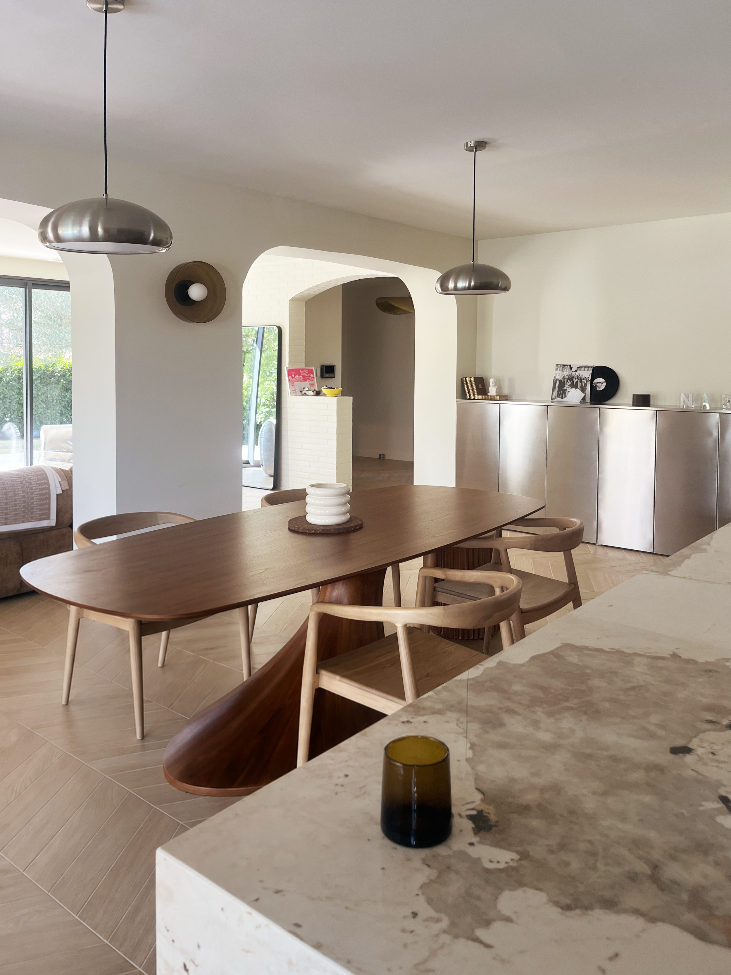 Intérieur moderne d'une salle à manger avec une table en bois, plusieurs chaises en bois, un plateau avec une jarre blanche, et une fontaine en verre teinté foncé. En arrière-plan, des lampes suspendues en métal, un mur blanc avec des décorations et une pièce lumineuse avec de grandes fenêtres.