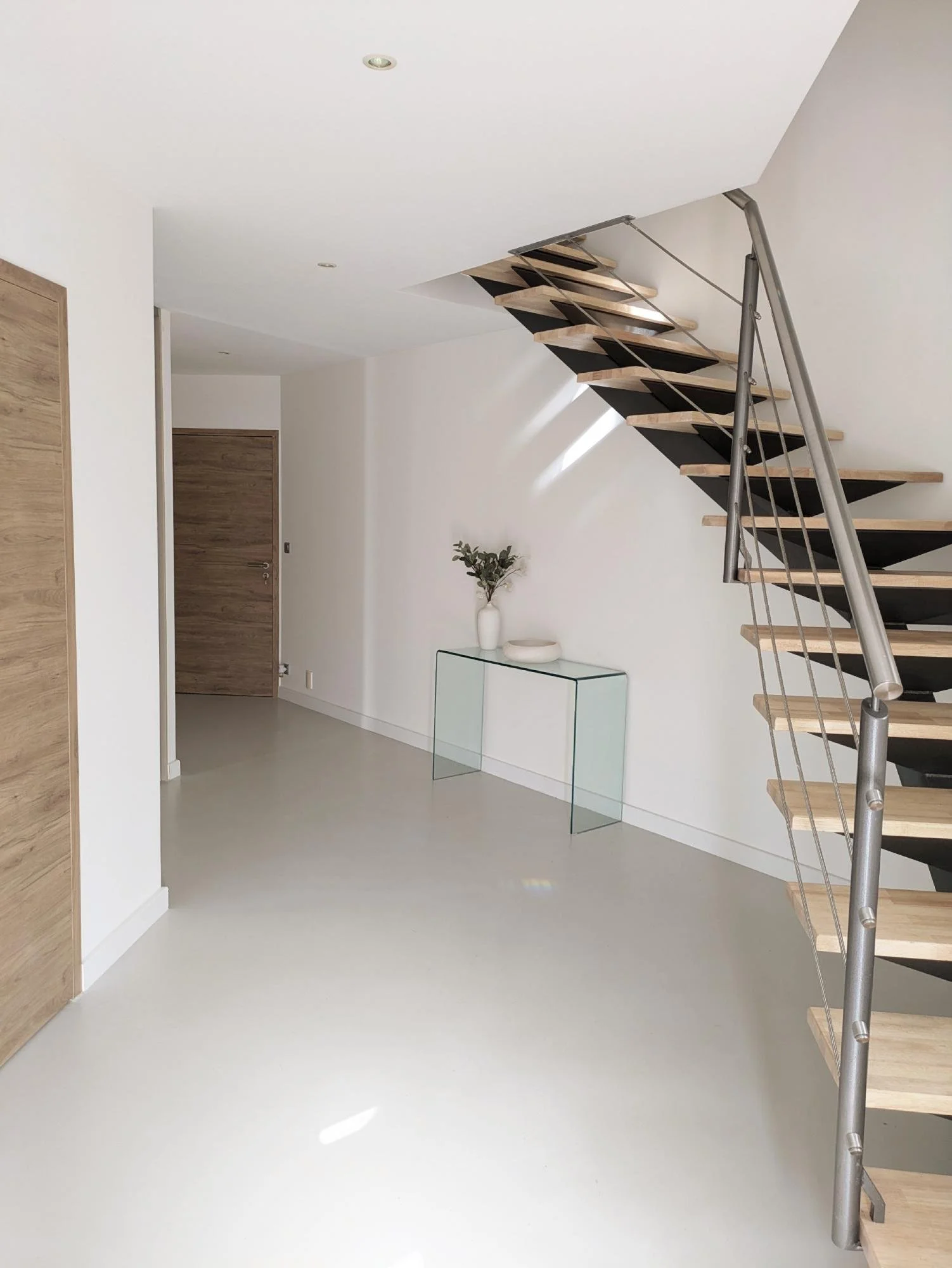 Intérieur moderne avec escalier en bois et métal, vase avec des feuilles vertes sur une console en verre, mur blanc, porte en bois, sol clair.