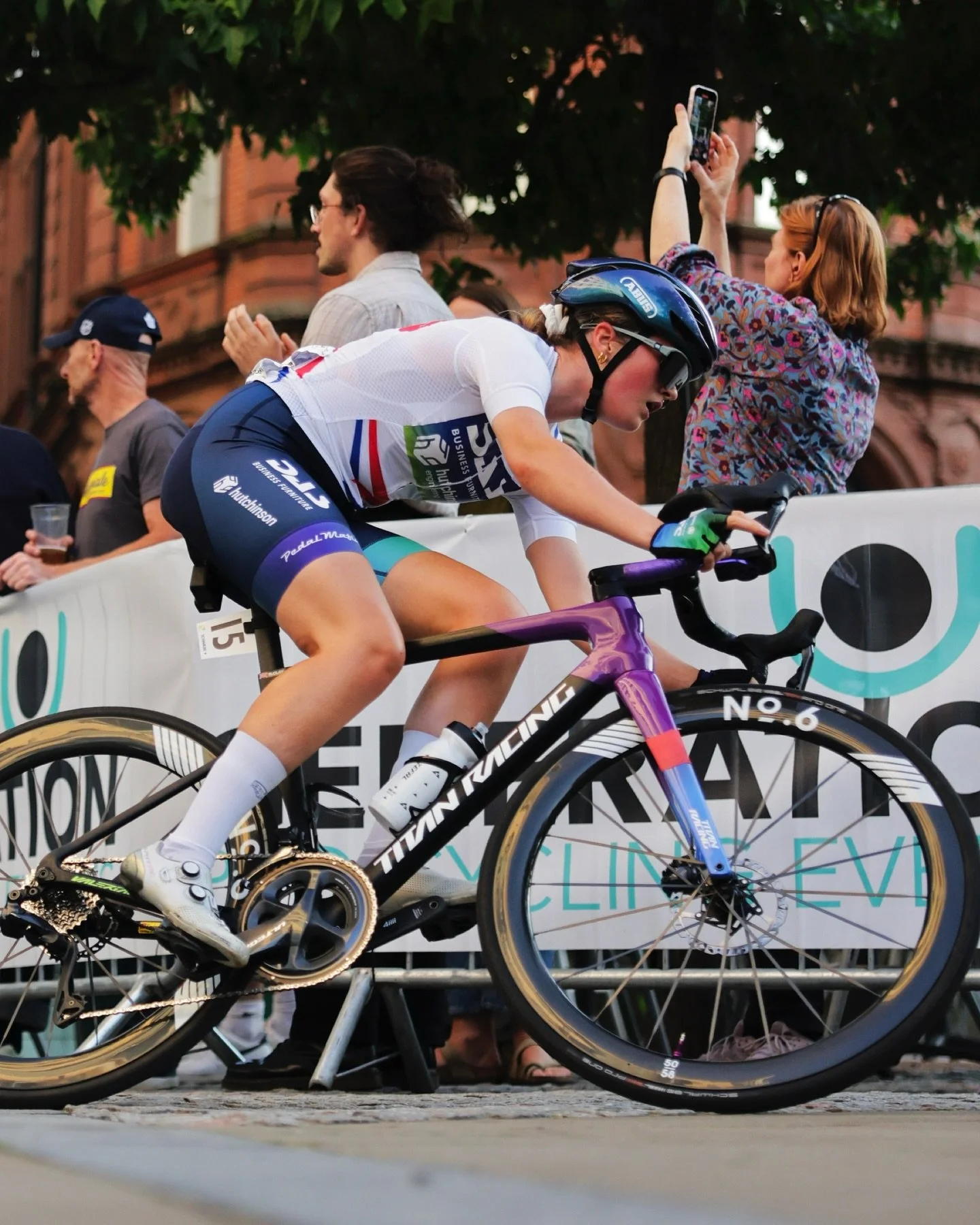 A female cyclist in a white jersey and blue shorts riding a purple racing bike during a race. She is wearing a helmet and sunglasses, and appears focused. Spectators watch behind a white barrier, with one woman taking a photo with her phone.
