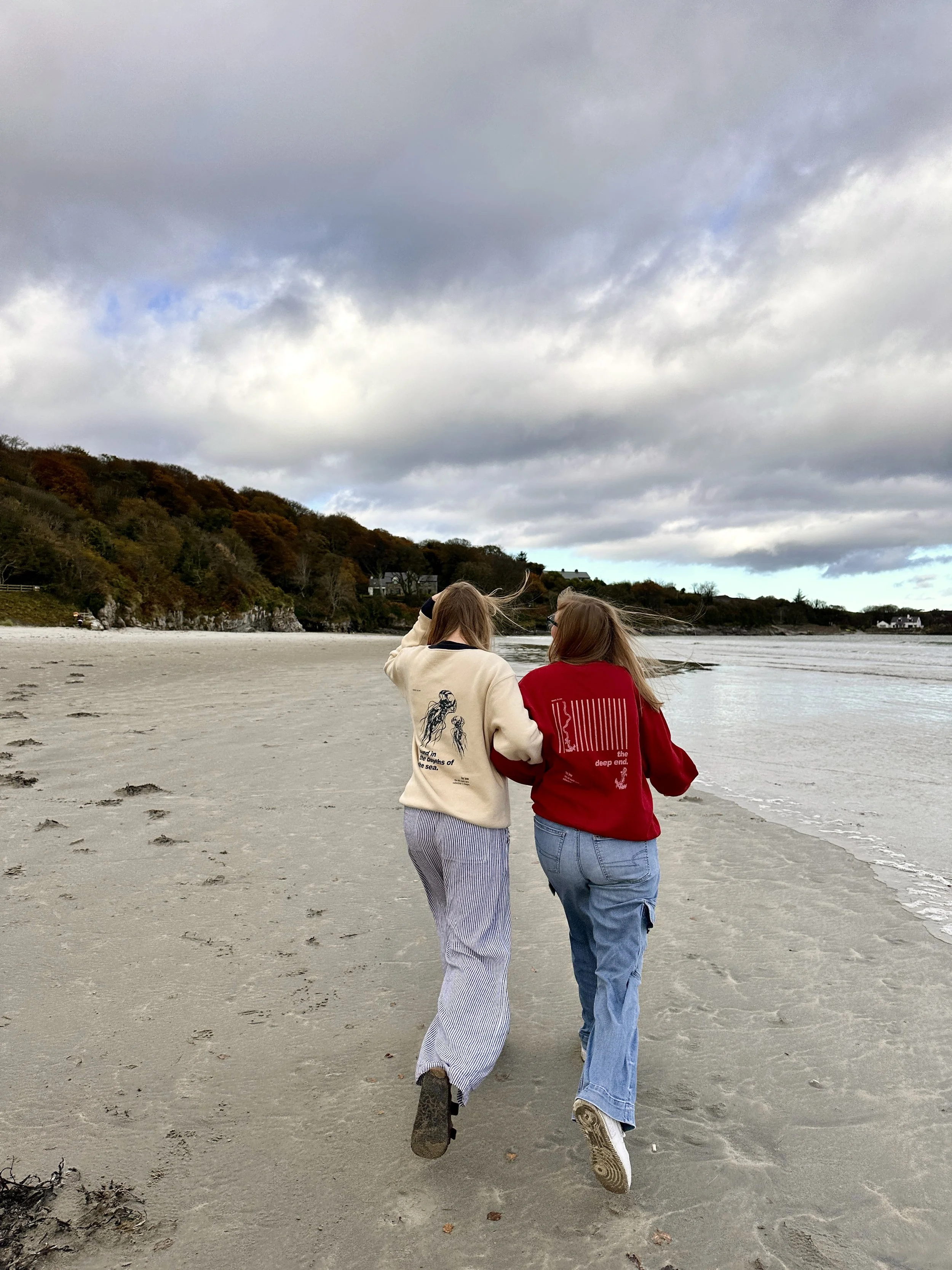 Two young women walk arm in arm along a sandy beach under cloudy skies. One wears a cream-colored jacket with a graphic, and the other wears a red jacket with text and bar code graphics. There are hills with trees in the background.