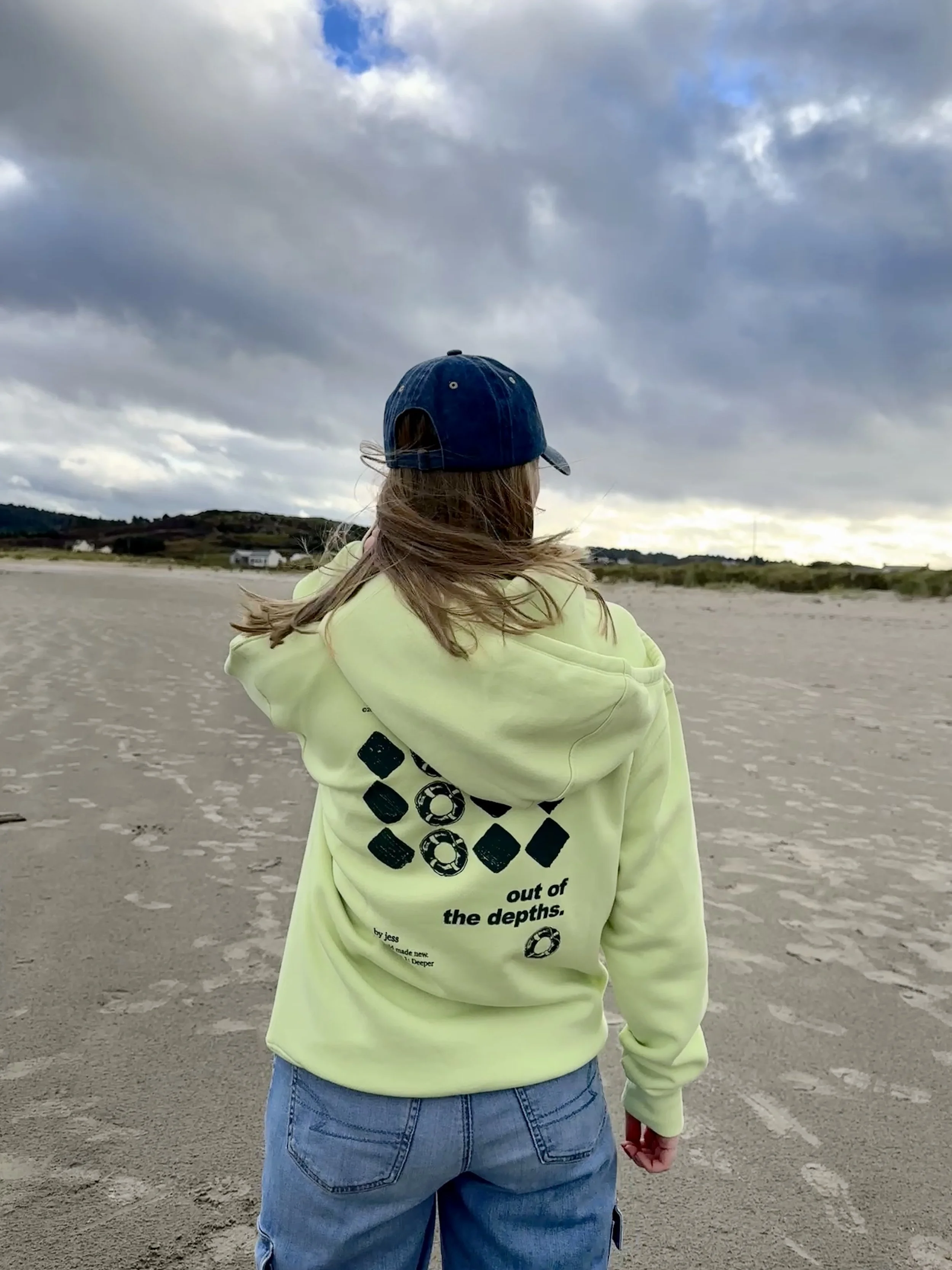 A woman with long hair wearing a navy baseball cap, a bright yellow hoodie with black graphic designs on the back, and blue jeans is standing on a sandy beach under a cloudy sky, looking towards the horizon.