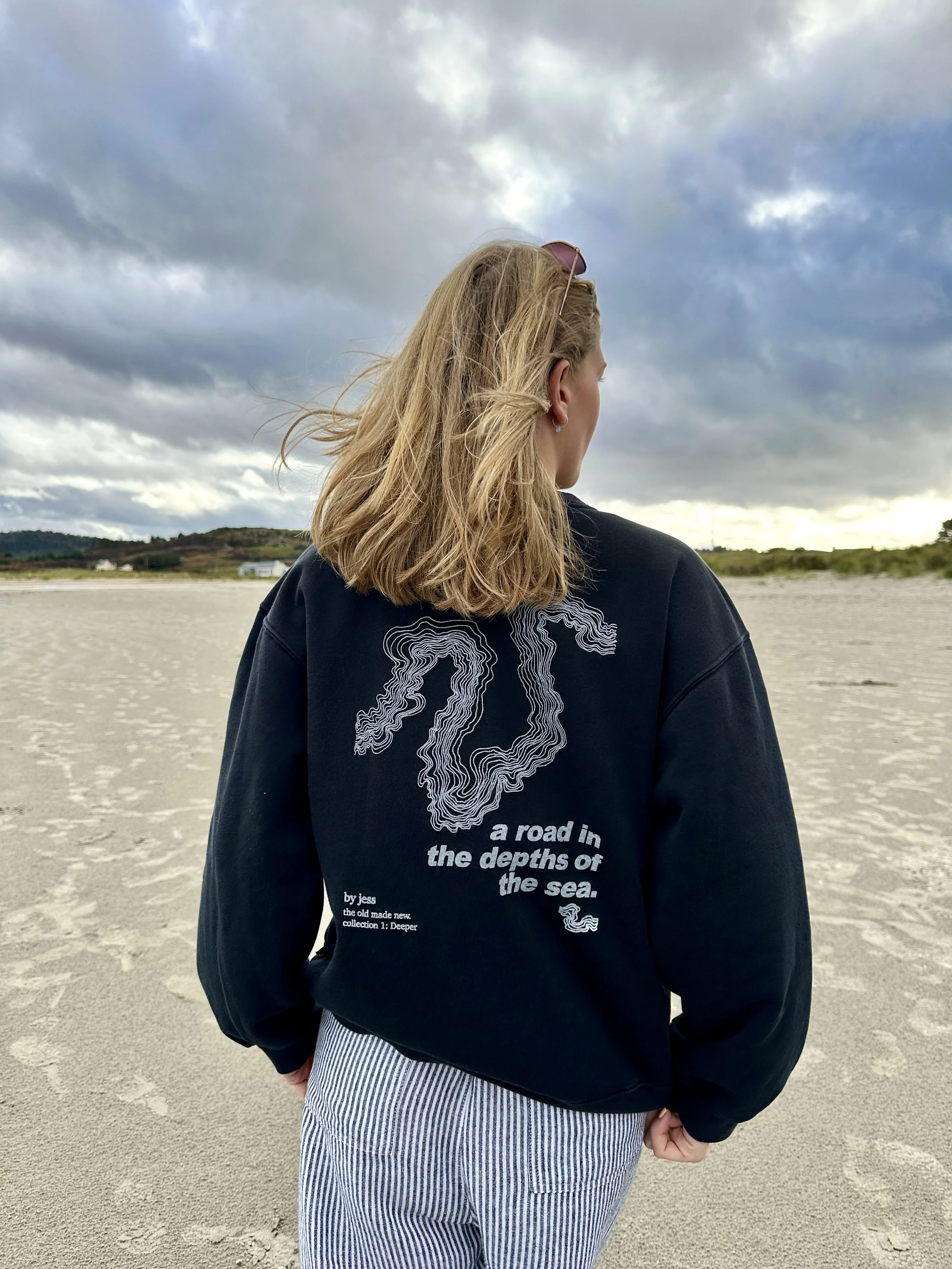 Woman with blonde hair wearing a navy sweatshirt with wave design and text, standing on a sandy beach under cloudy sky.