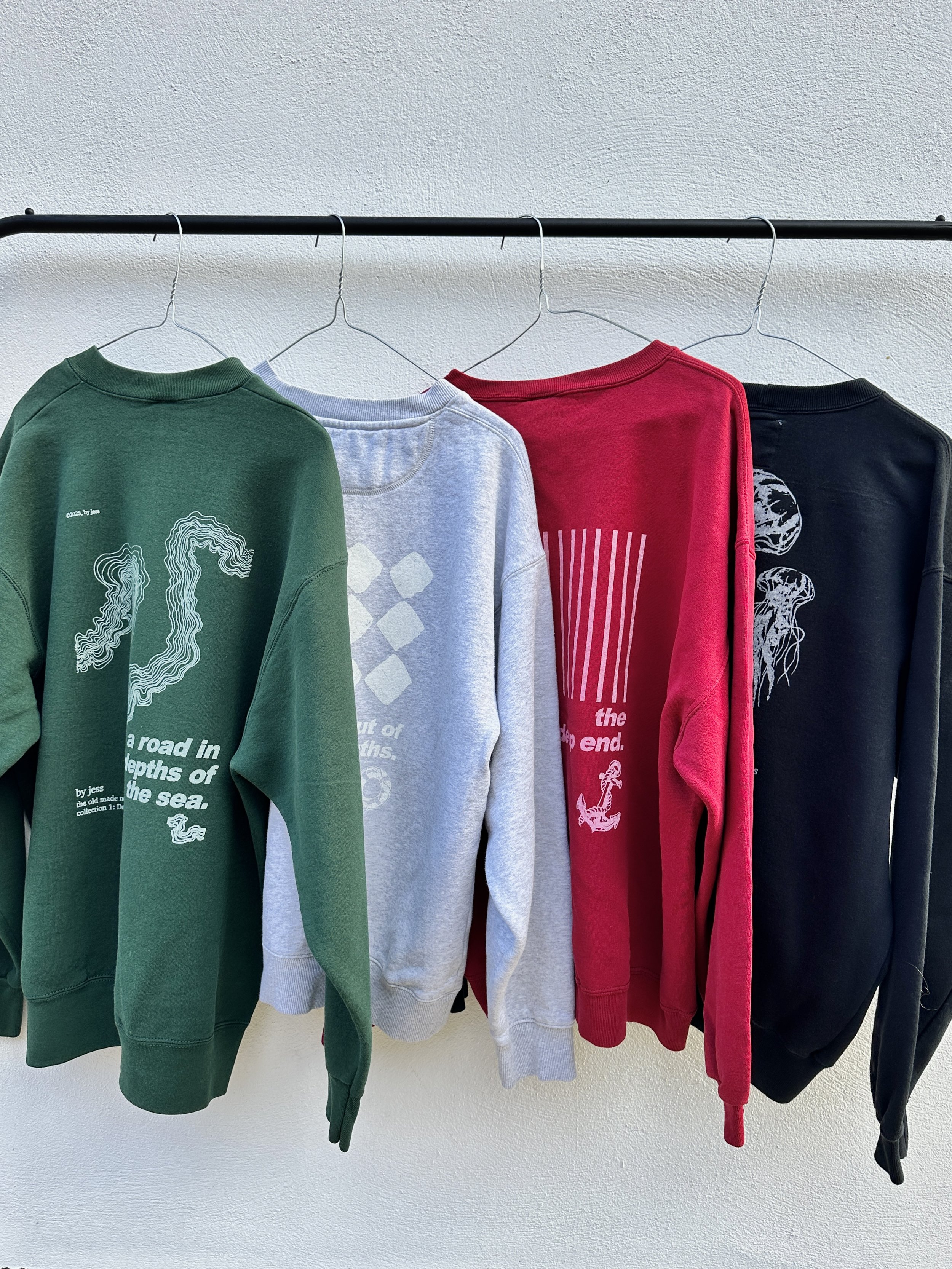 Four sweatshirts hanging on a clothing rack against a white wall, in green, light gray, red, and black colors, with nautical and abstract designs.