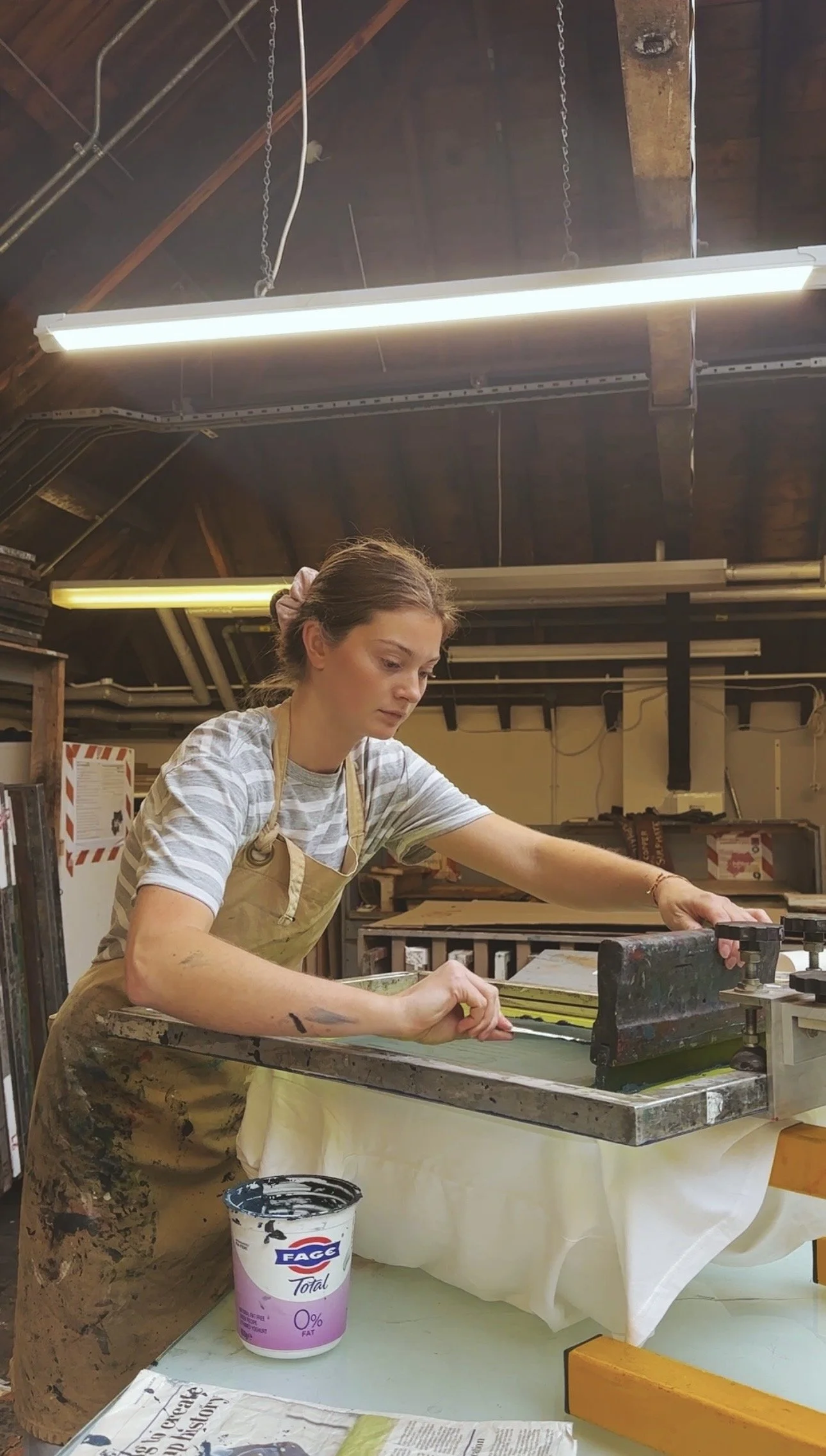 A woman working in a woodworking shop, wearing a striped t-shirt and an apron, smoothing a piece of material on a workbench with a tool, with paint or glue on her arms, and a bucket of face total zero fat cream nearby.