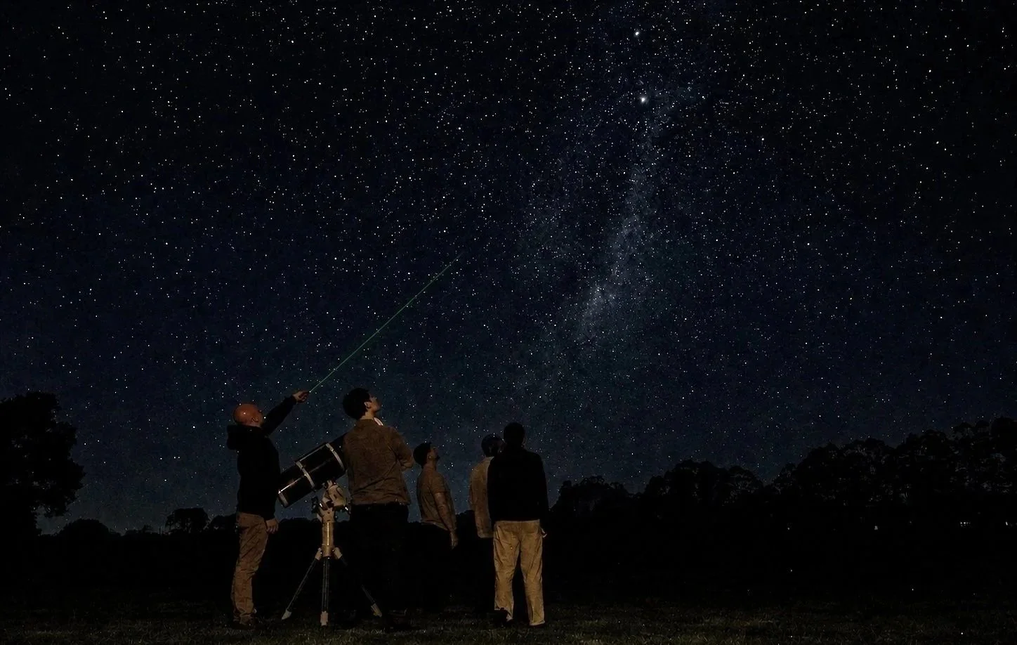 Group of people stargazing at night with a telescope, looking at the star-filled sky and the Milky Way.