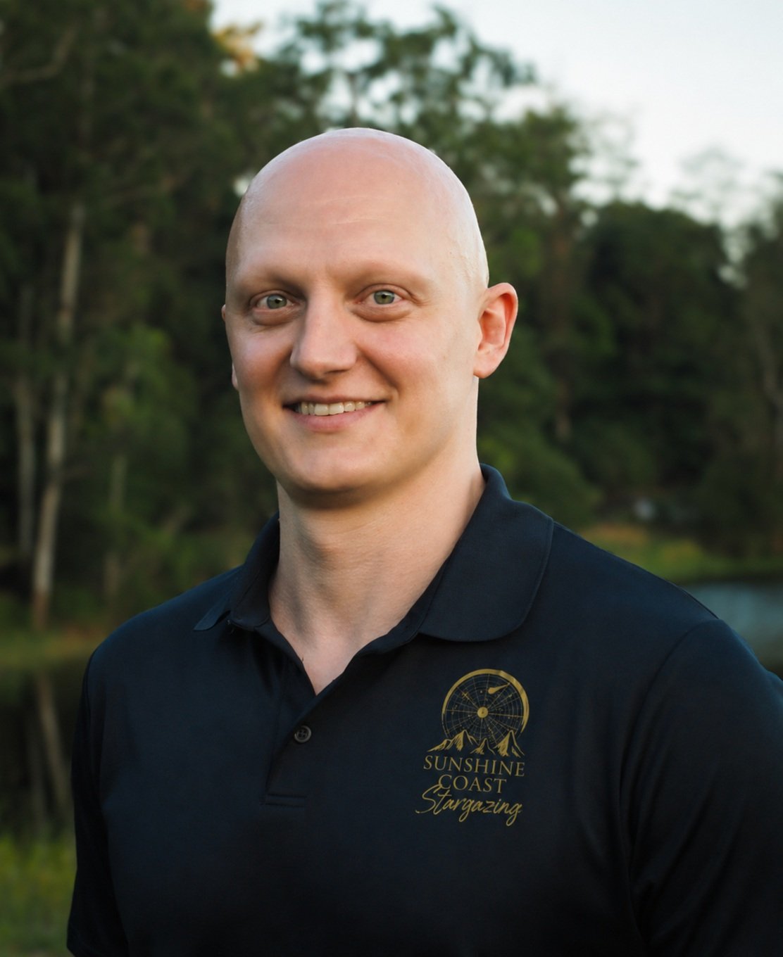 A man smiling outdoors, wearing a black polo shirt with a logo that says "Sunshine Coast Stargazing," in front of a background of trees and a body of water.