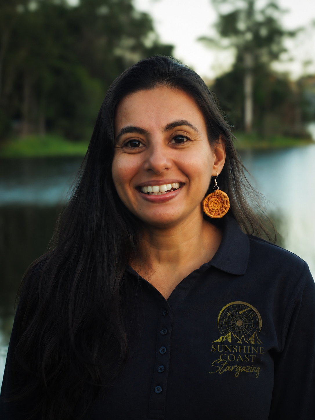 A woman smiling outdoors near a body of water, wearing a black polo shirt with a logo that says 'Sunshine Coast Stargazing' and an orange earring.