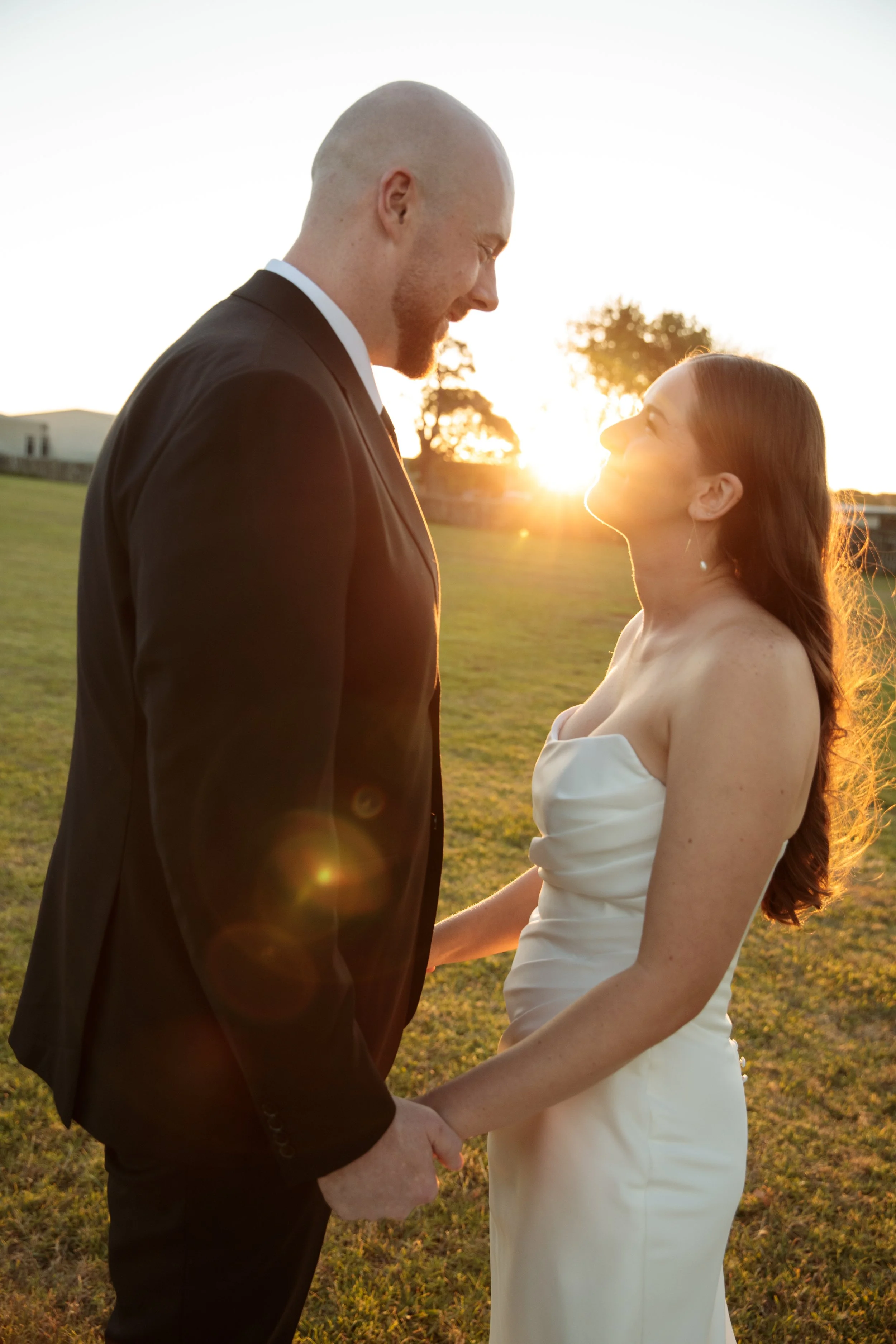A newlywed couple holding hands and gazing at each other in a grassy open field during sunset.