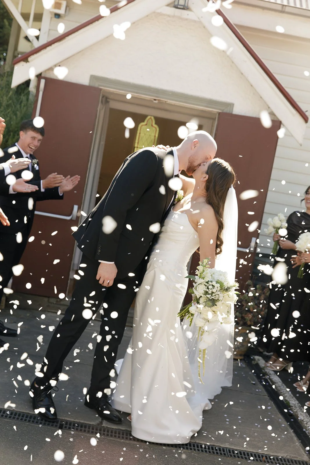 A newlywed couple sharing a kiss as confetti falls, surrounded by friends clapping and celebrating outside a house.