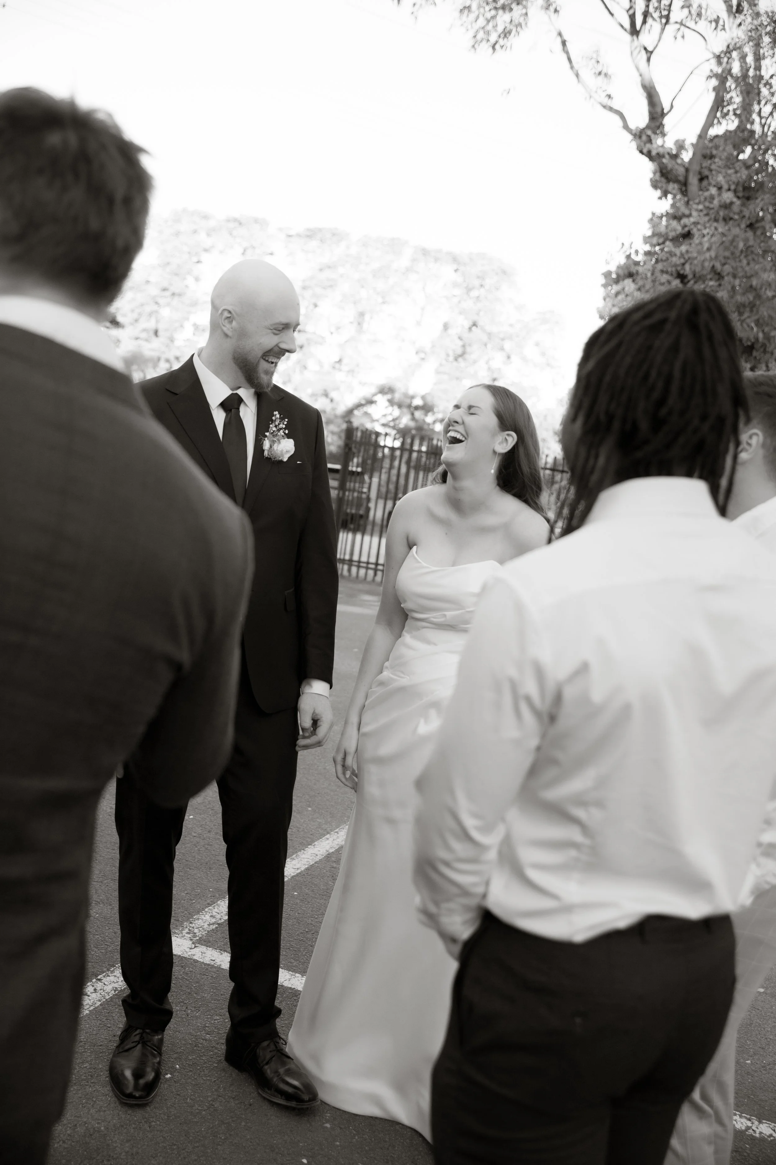 Black and white photo of a joyful bride and groom laughing together outdoors, surrounded by friends, during a wedding celebration.