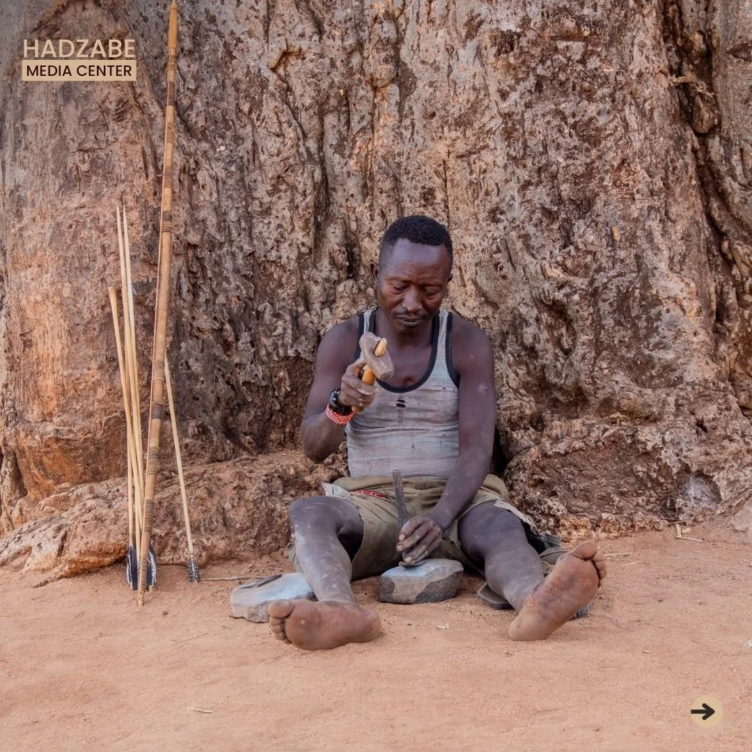 A picture showing a Hadzabe man sitting under a tree hammering at something whi